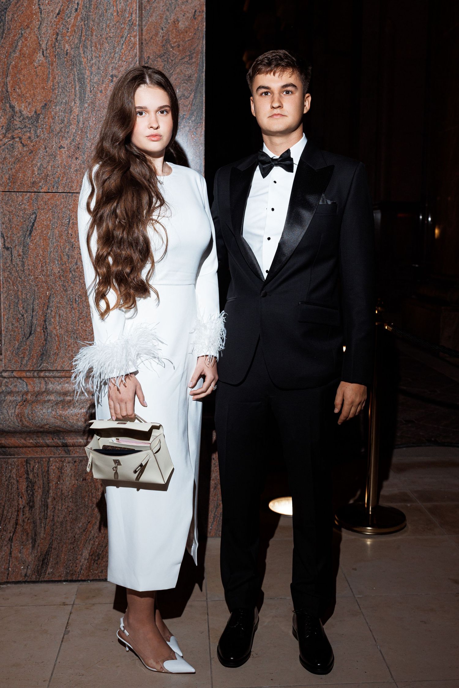 A couple in formal attire poses against a textured wall, with her in a white feathered dress and him in a black tuxedo.