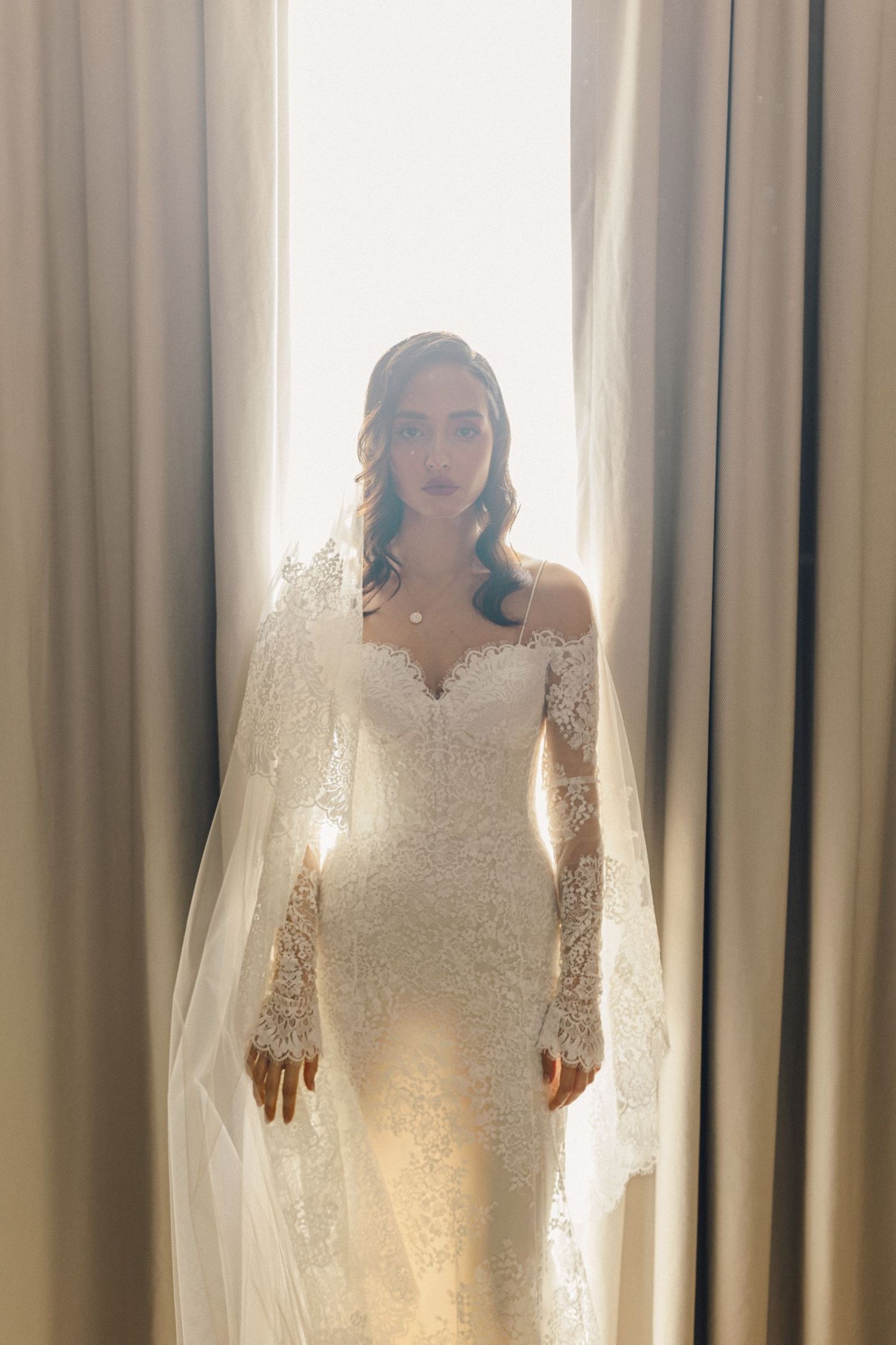 Sunlit bridal portrait in front of curtains showing off a long-sleeved lace wedding dress.