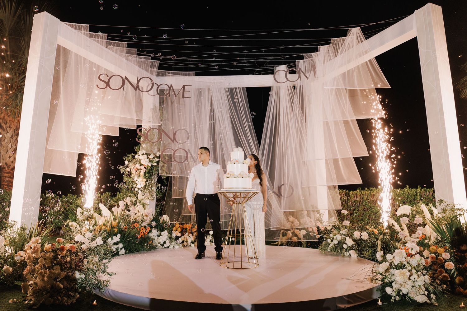 White draped wedding ceremony space decorated with floral arrangements and hanging lights.