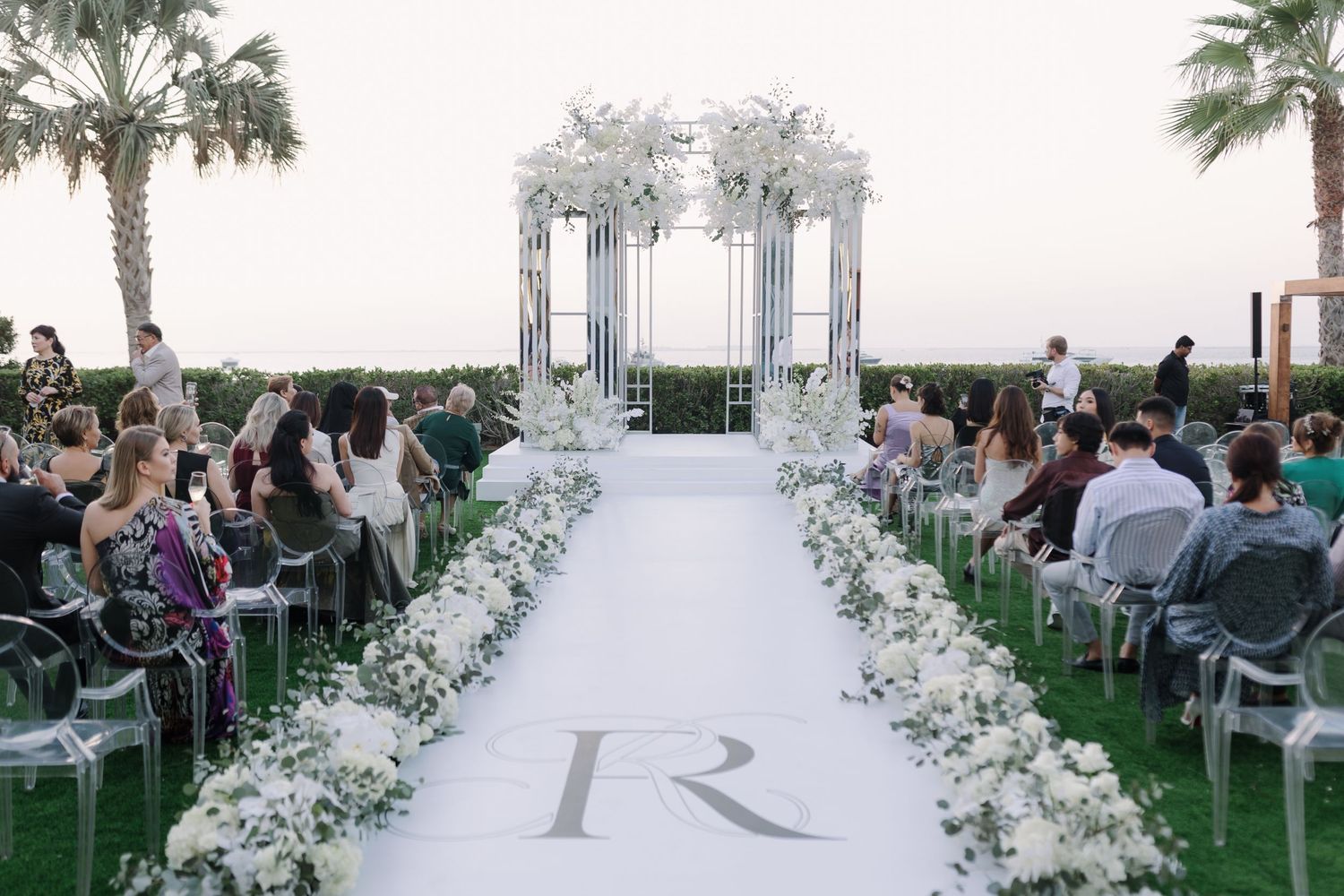 Elegant outdoor wedding ceremony setup with white floral decorations and clear chairs.
