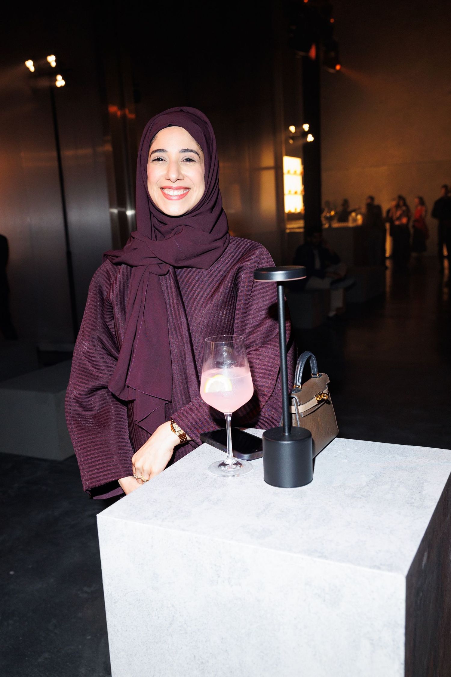 Woman in purple hijab smiling with cocktail at modern event space.