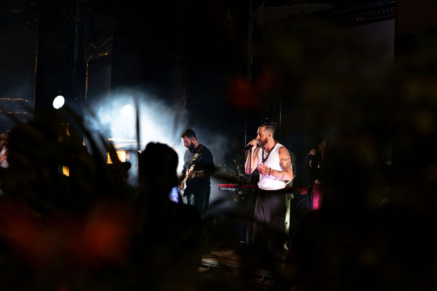 Musicians perform on a dark stage with dramatic lighting and silhouetted audience members in the foreground.