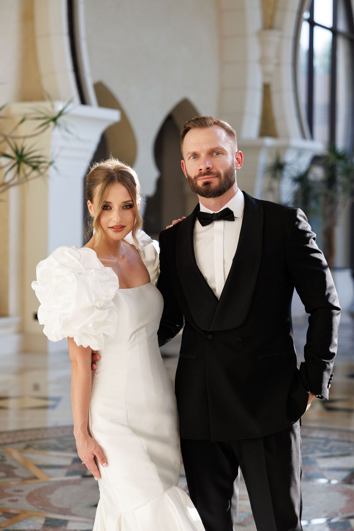 A bride in white and groom in black tuxedo pose together in an elegant indoor setting.
