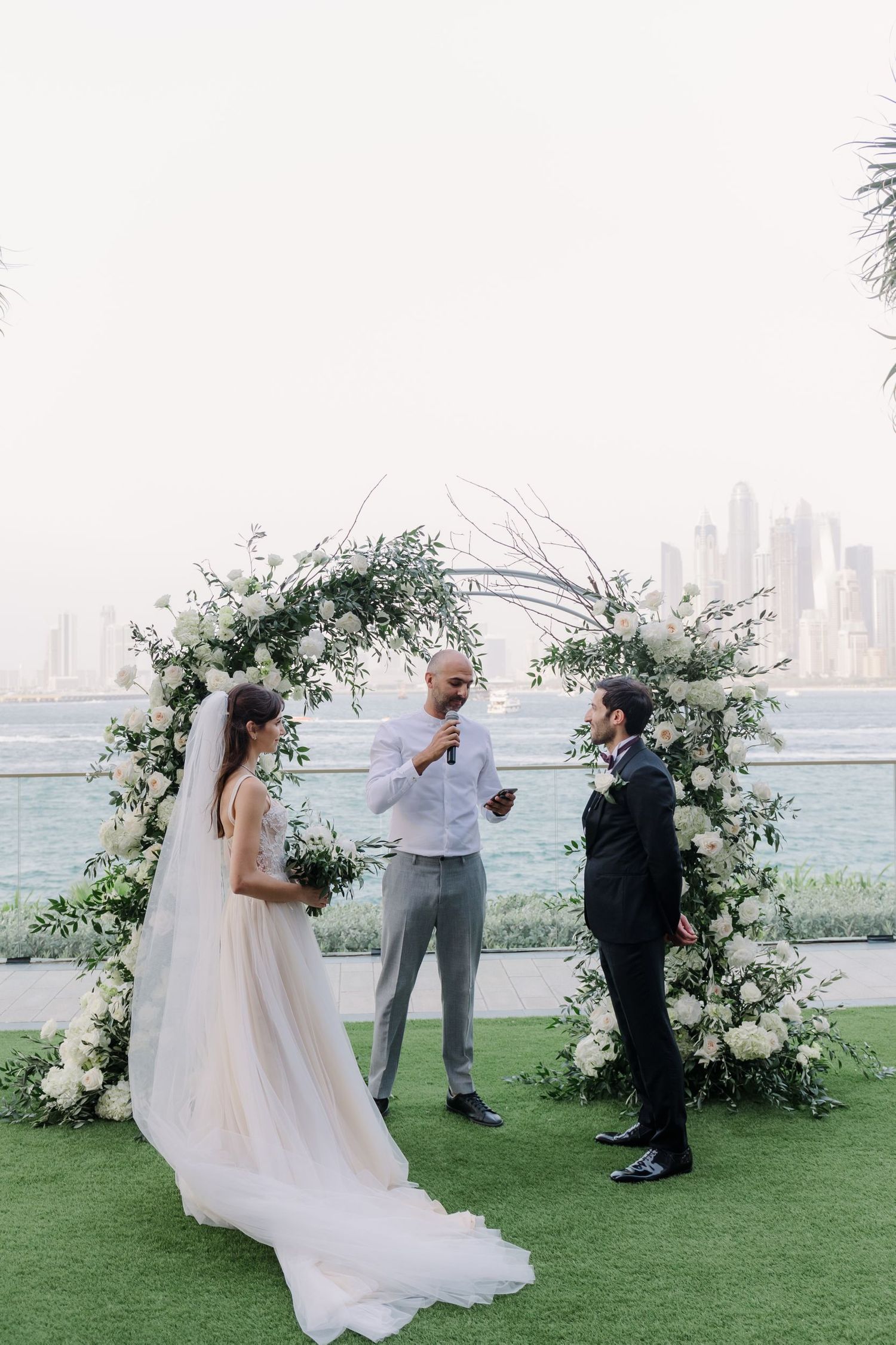 Wedding ceremony setup with floral arch and city skyline backdrop on a grassy lawn.