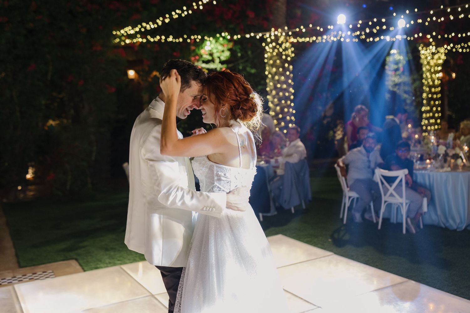 Couple dancing at night under blue spotlights and string lights at outdoor wedding reception.