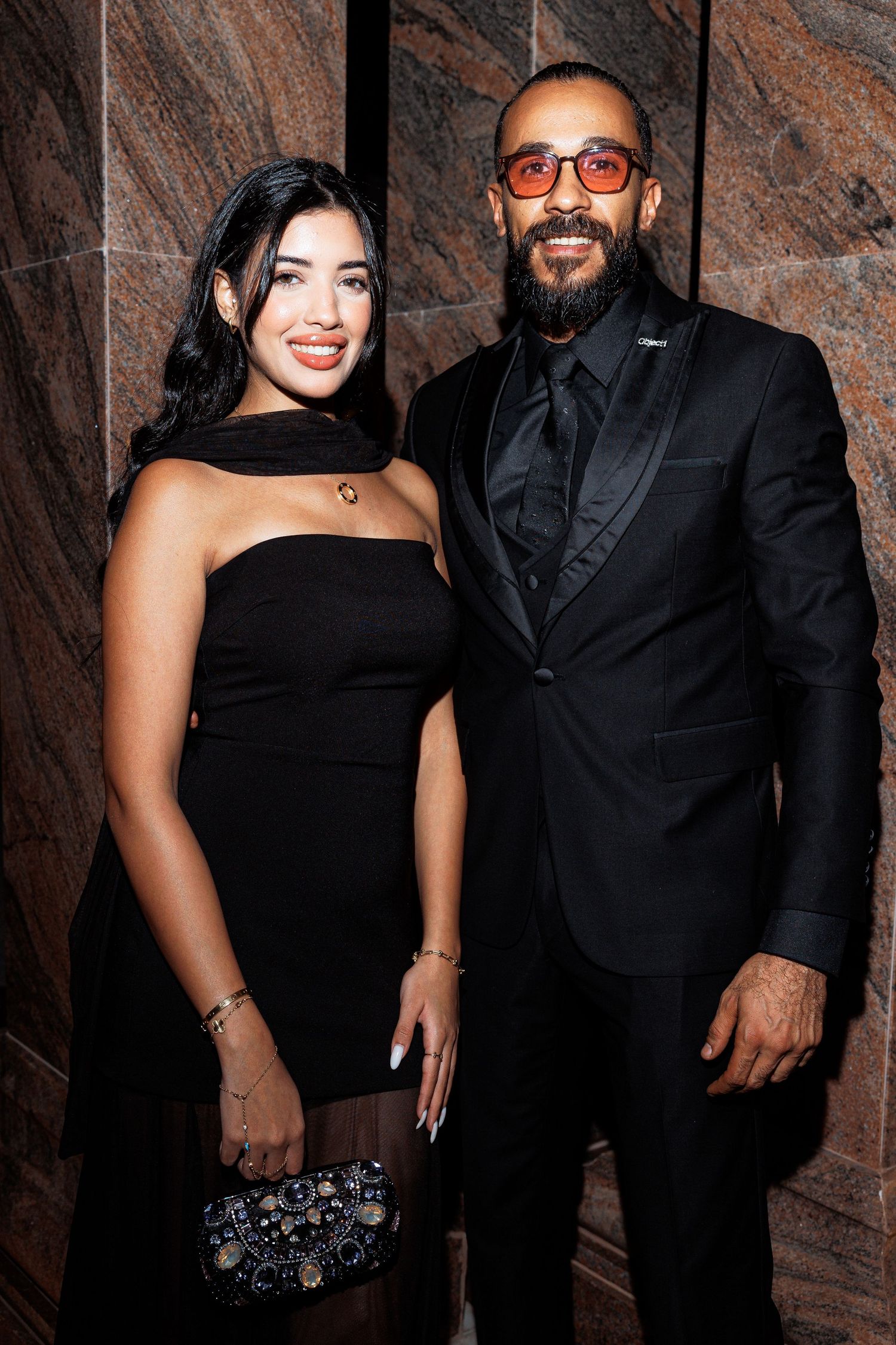 Two people in formal black attire pose together against a textured stone wall at an evening event.