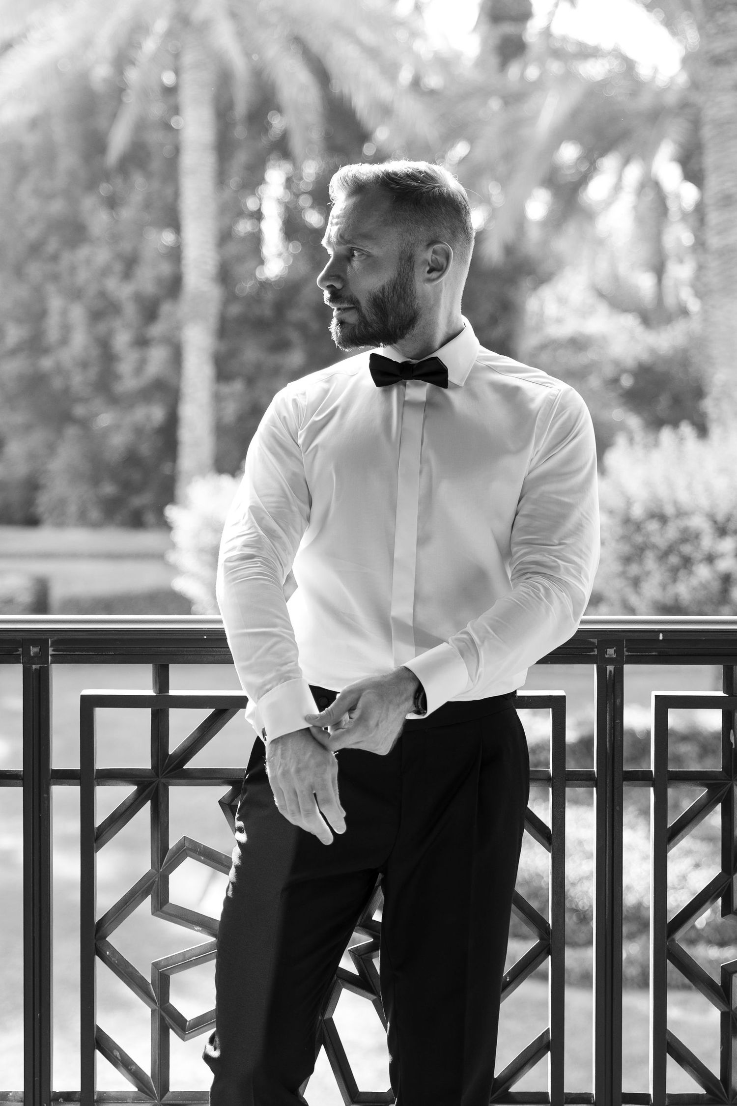 Black and white photo of groom in white shirt and bowtie sitting on railing.