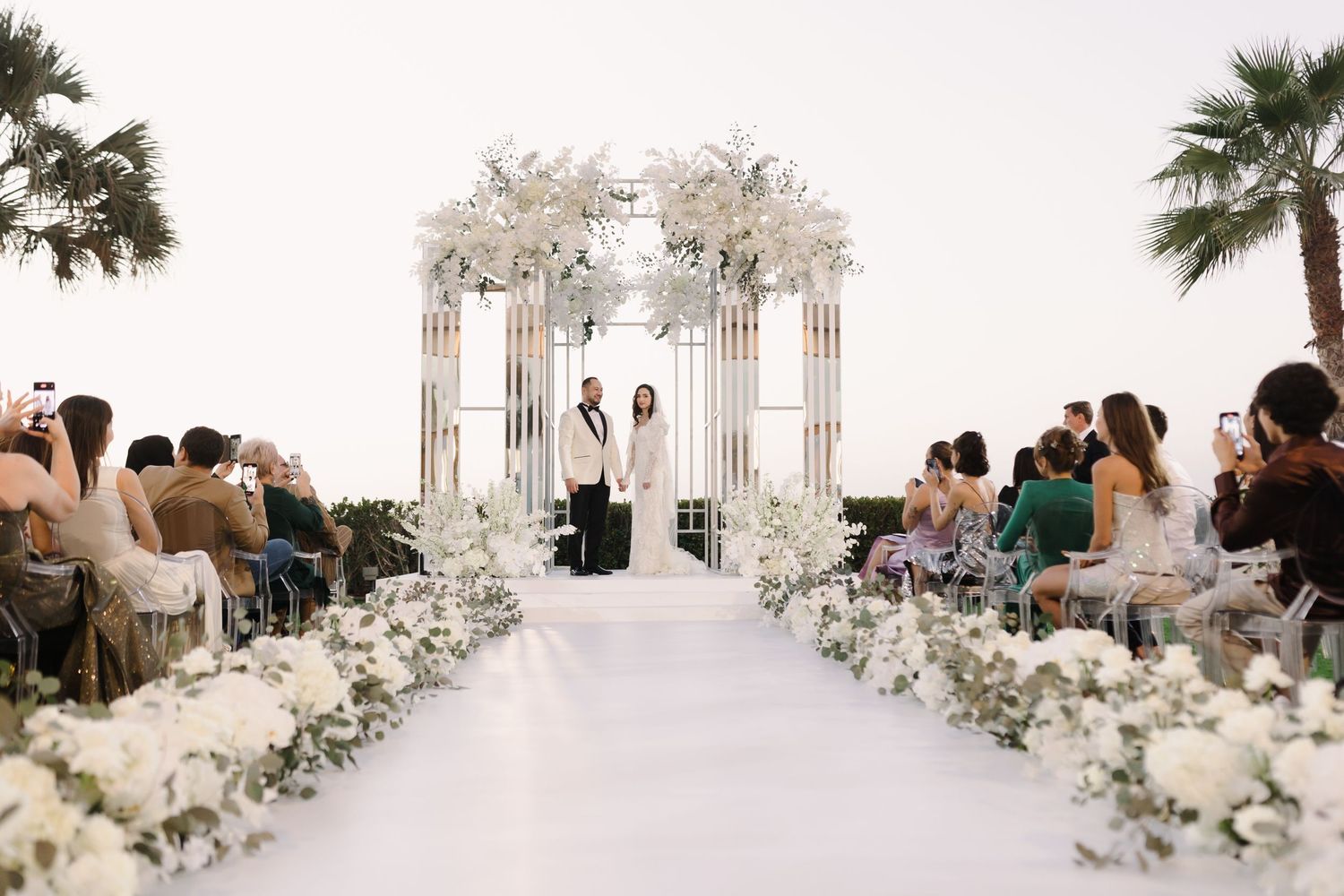 An elegant wedding ceremony setup with white floral arches and chairs under palm trees.