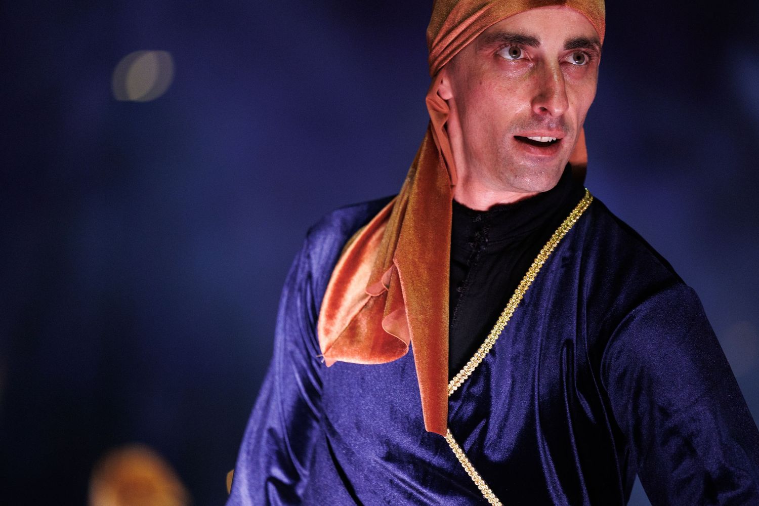 Performer in navy costume with orange headwrap against dark blue stage lighting.