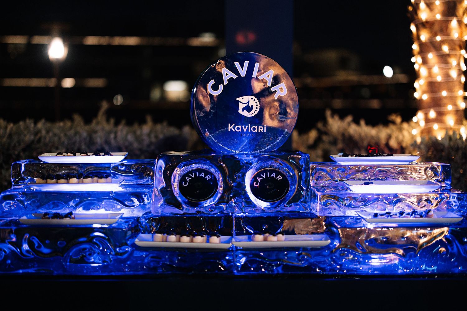 A blue-lit ice sculpture display featuring KAVI vodka bottles and glasses arranged on an illuminated bar setup.