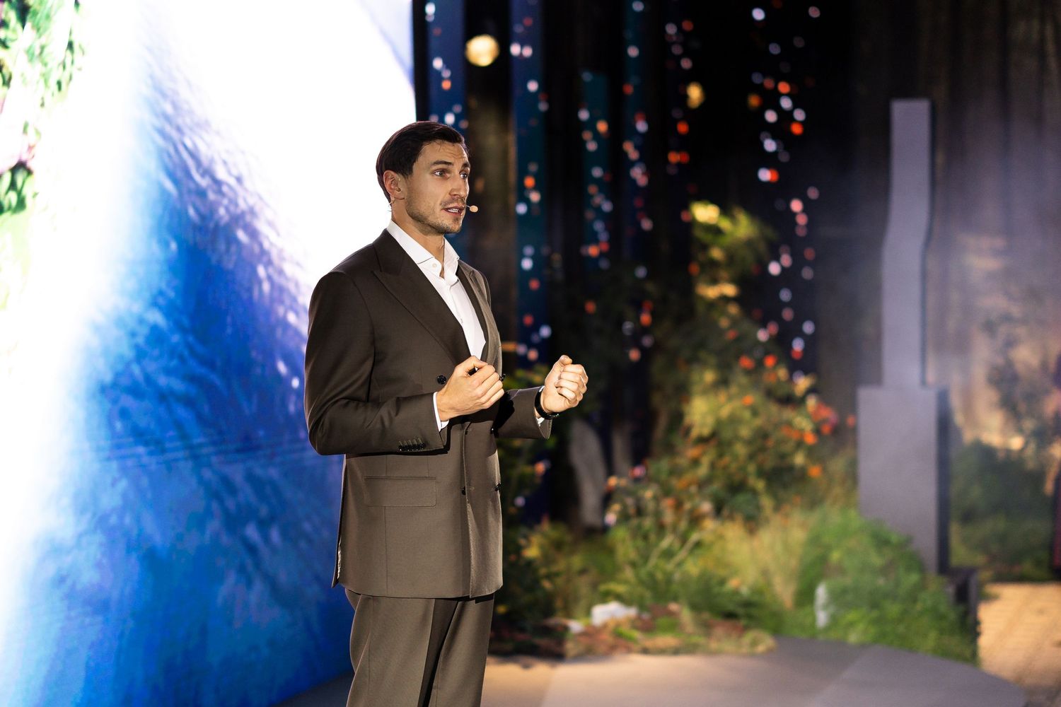 A speaker in a dark suit gestures while presenting on stage with colorful lights in the background.