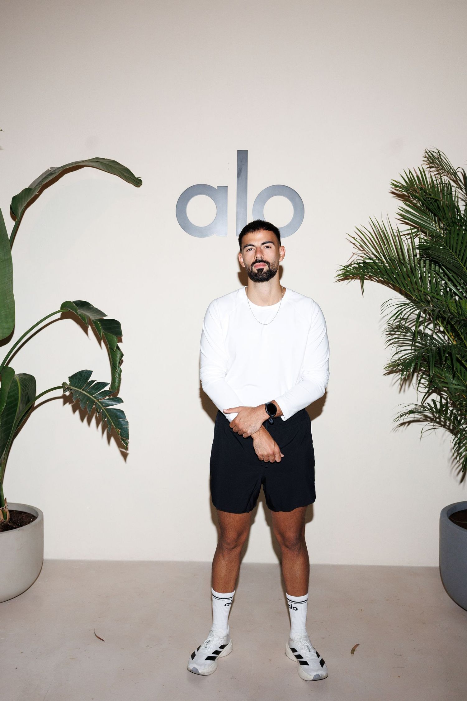 Person in white shirt and black shorts standing against white wall with alo logo and plants.