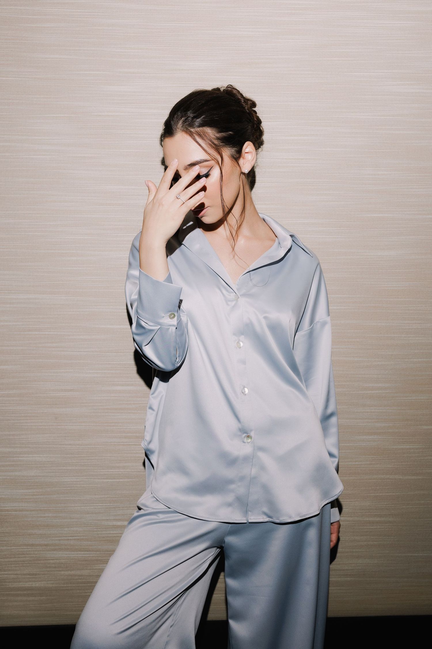 A person in light blue silk pajamas covering their face with one hand against a beige background.