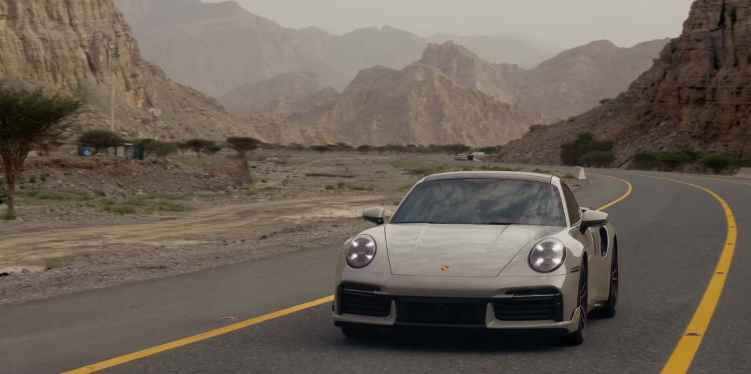 A silver Porsche driving on a desert road with dramatic mountain scenery.