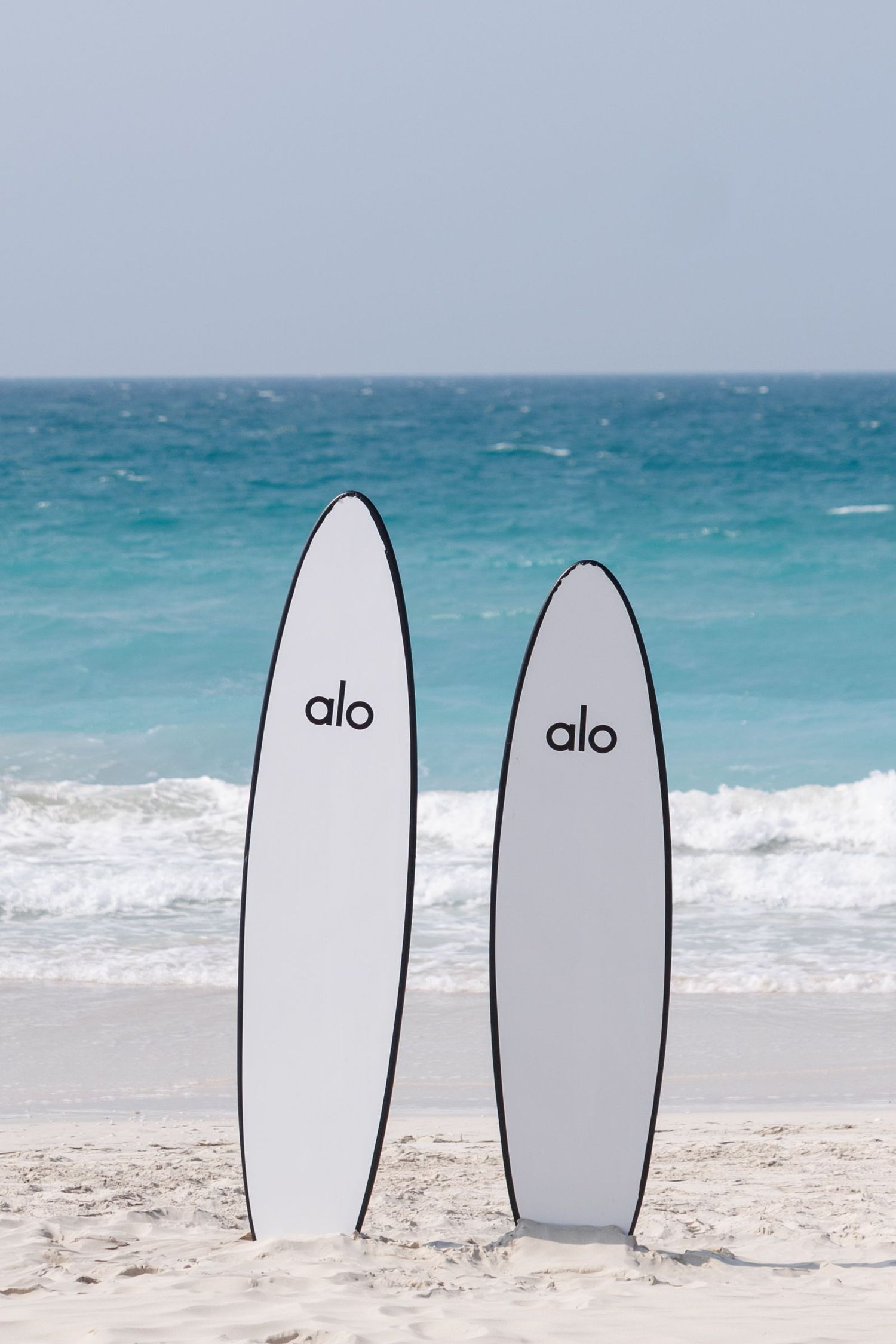 Two white Alo surfboards standing upright on pristine beach with turquoise ocean waves.