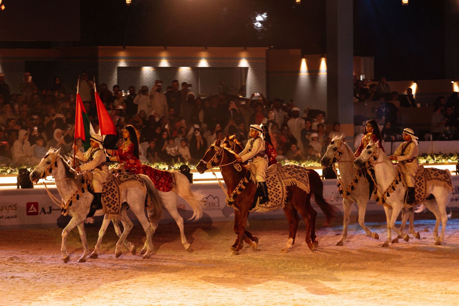 Performers in traditional costumes ride camels in a choreographed nighttime desert show with dramatic lighting.