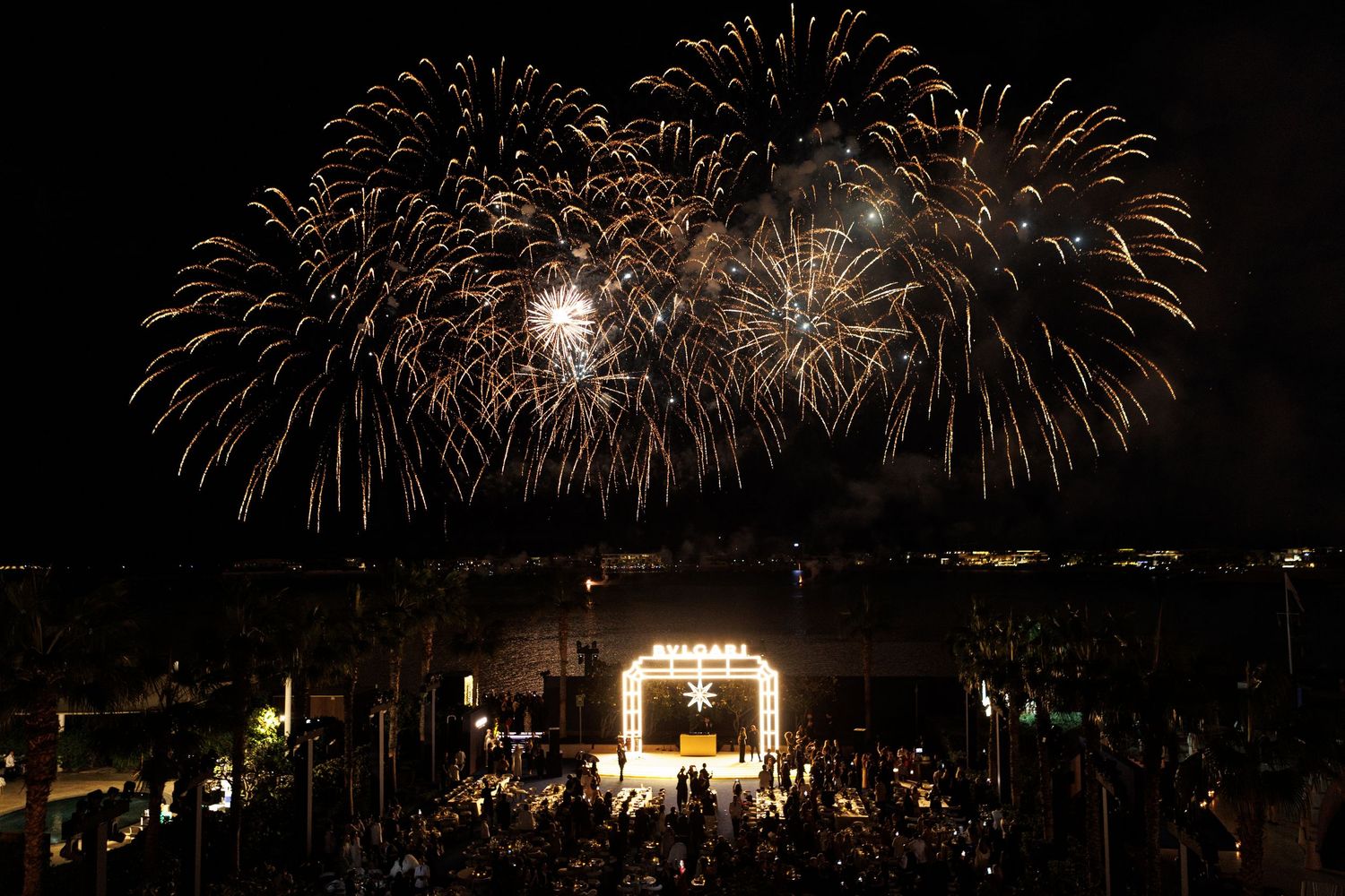 Spectacular fireworks display illuminates the night sky over a dark waterfront cityscape with multiple bursts of light.