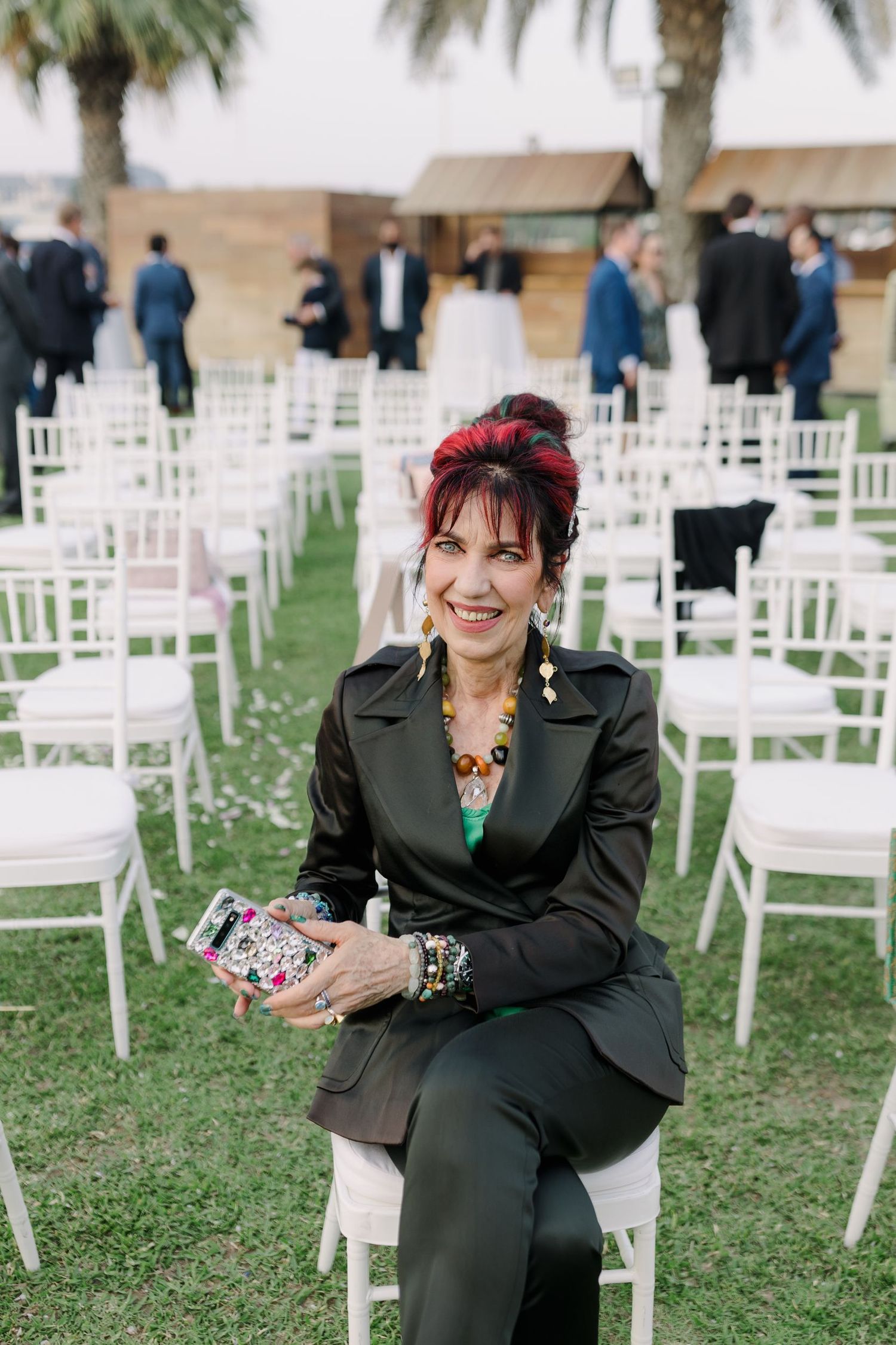 Guest seated among white chairs at outdoor wedding venue wearing dark suit and statement jewelry.