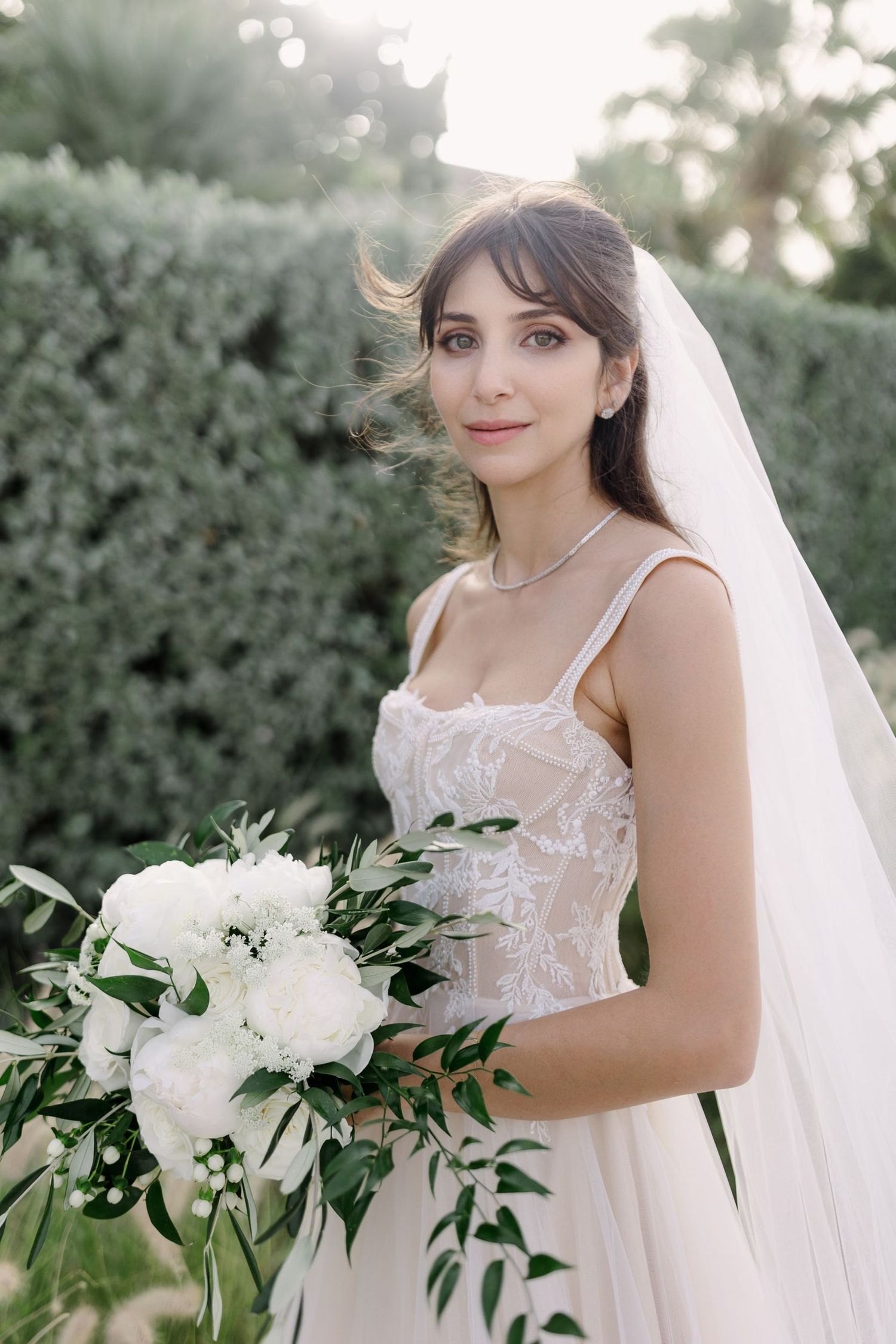 Bride holds a lush white floral bouquet with greenery in an outdoor garden setting.