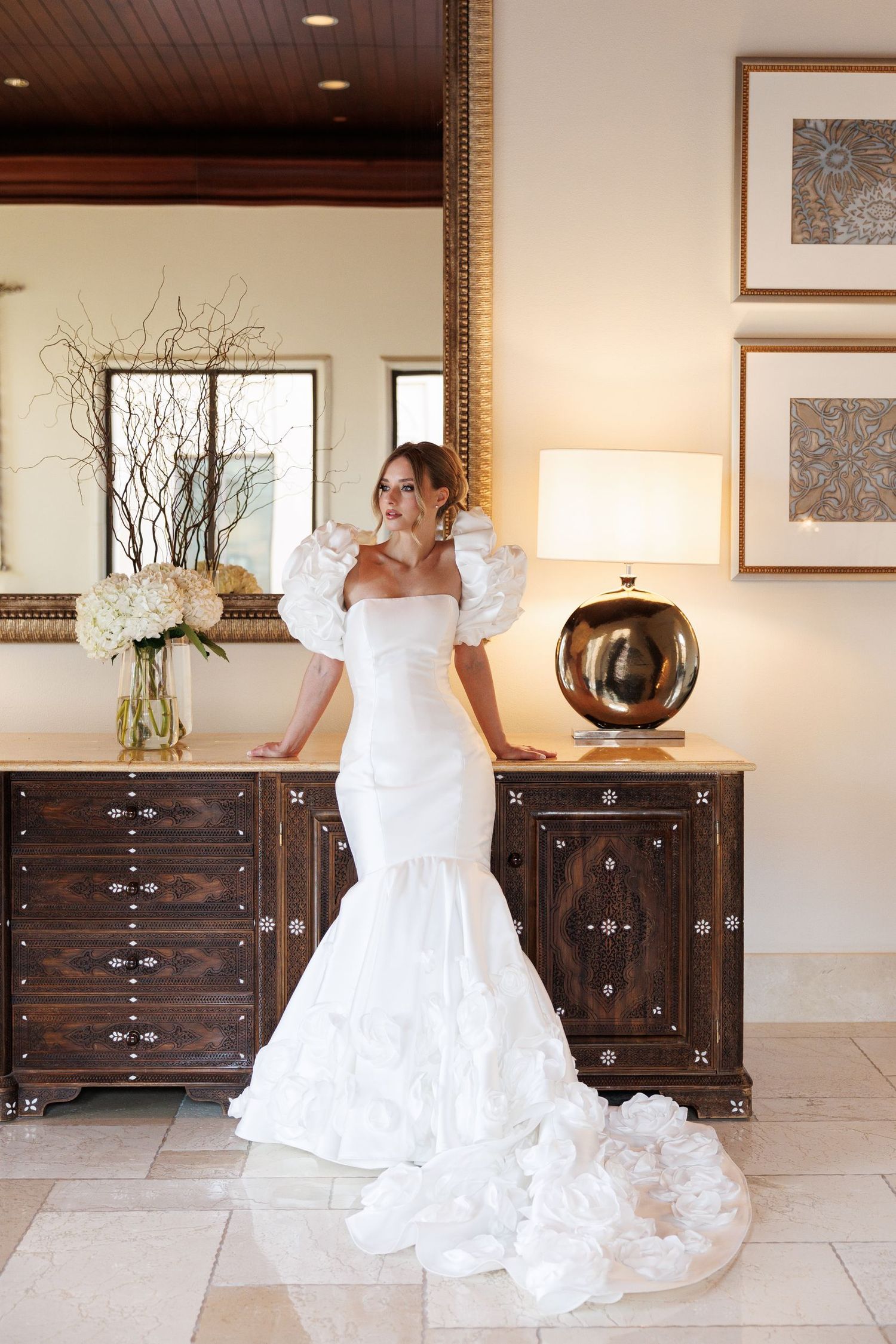 A bride in a flowing white gown poses dramatically against dark wooden furniture.