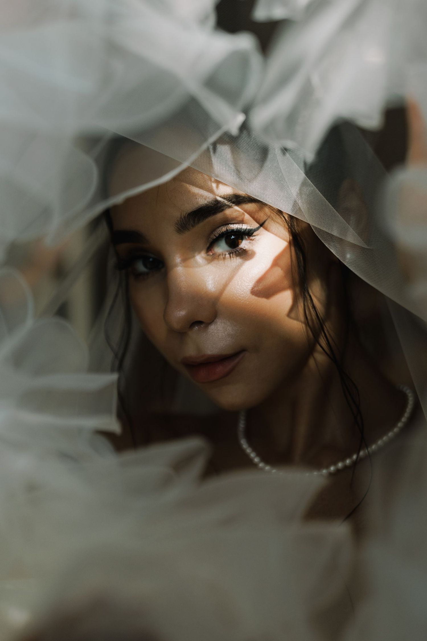 Close-up bridal portrait through sheer veil with pearl necklace and soft smile.