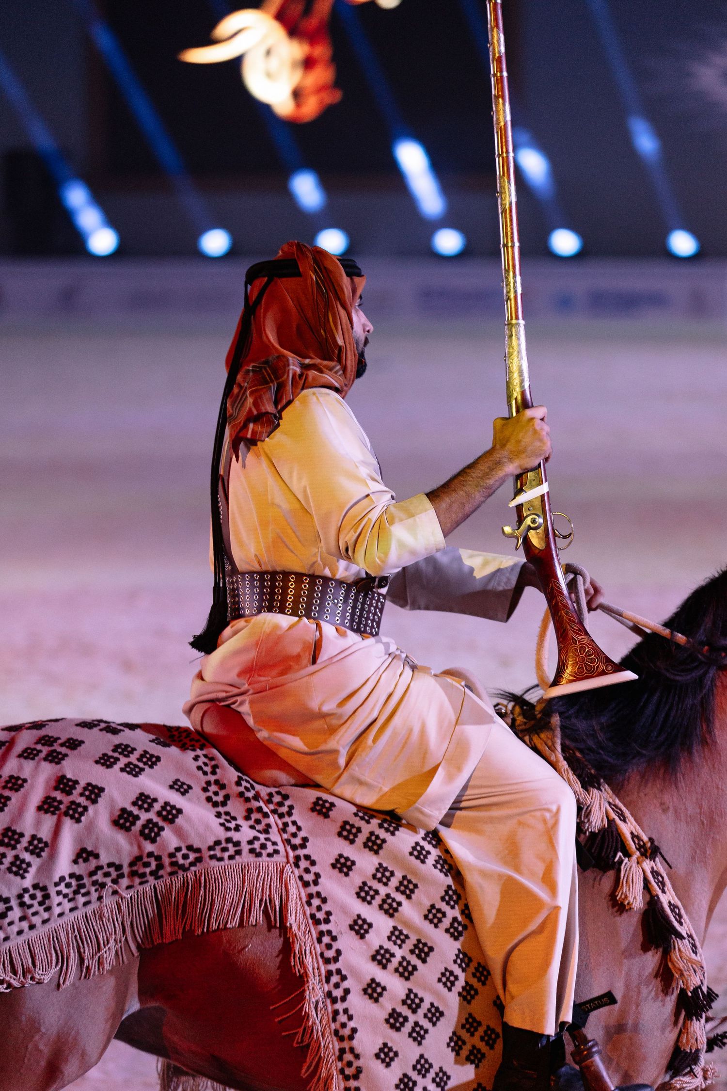 A rider in traditional white attire performs on horseback during an equestrian show with decorative patterns on display.