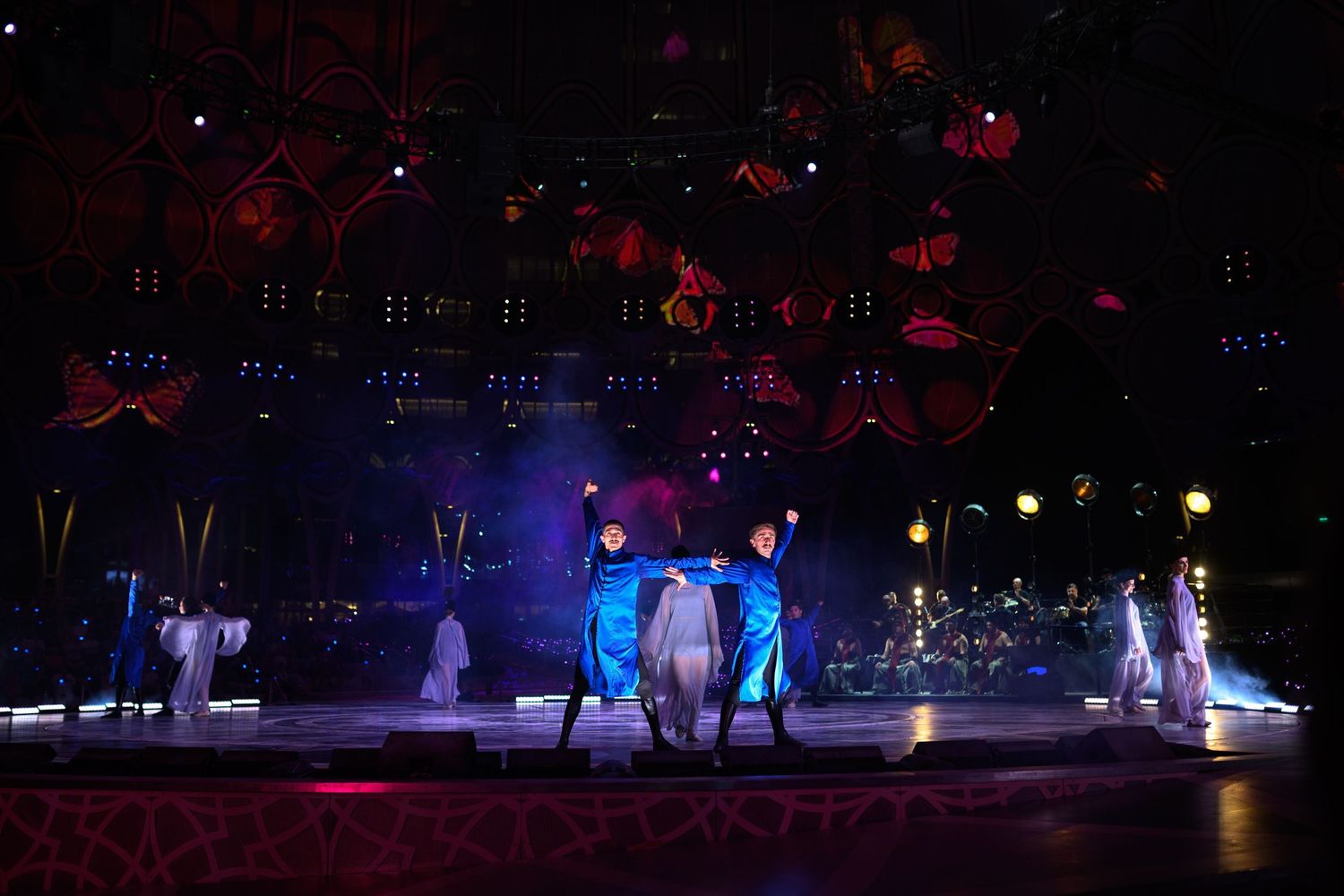 Stage performers in blue costumes under dramatic lighting with red decorative elements overhead.