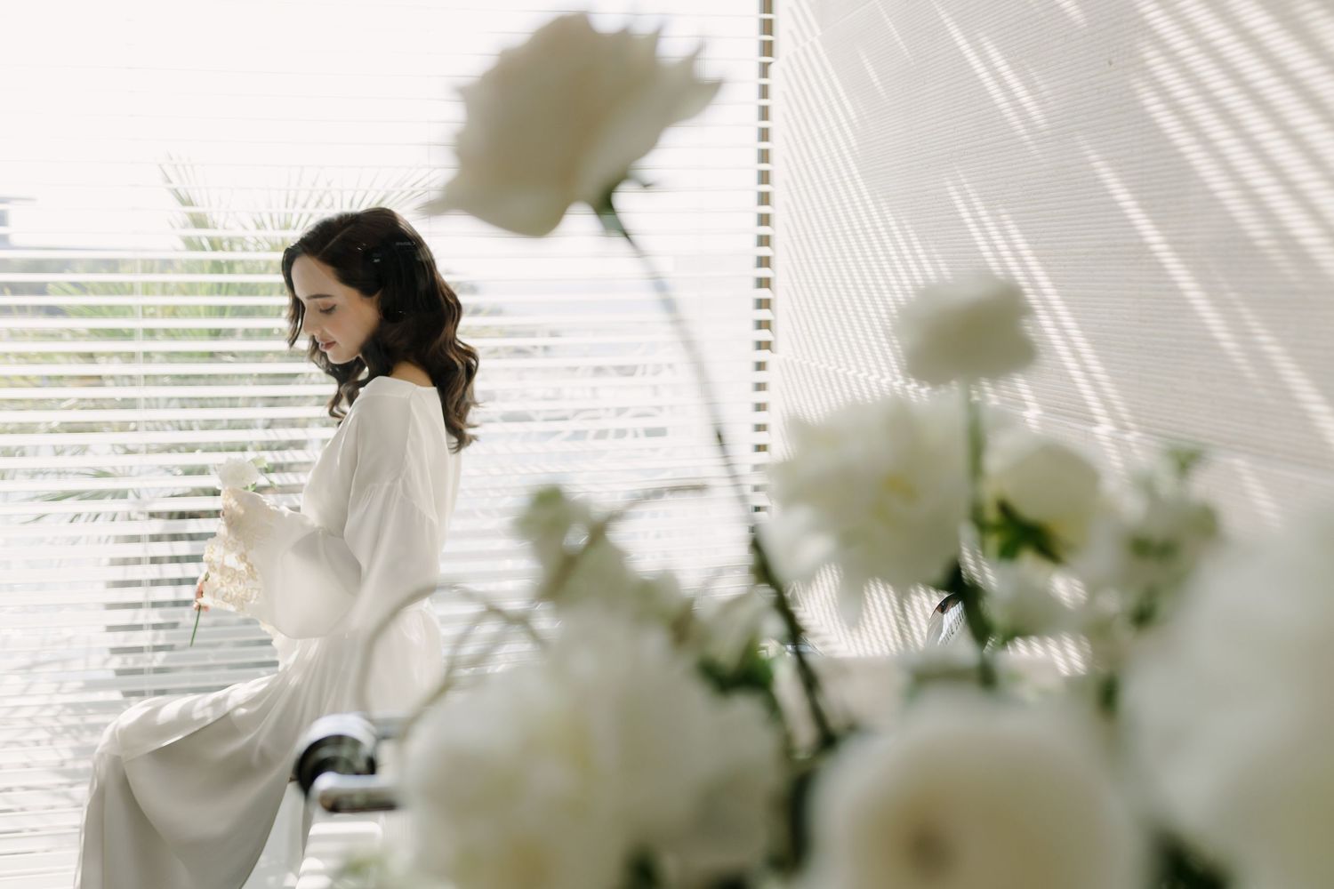 Ethereal portrait with white flowers and window blinds casting soft shadows in natural light.