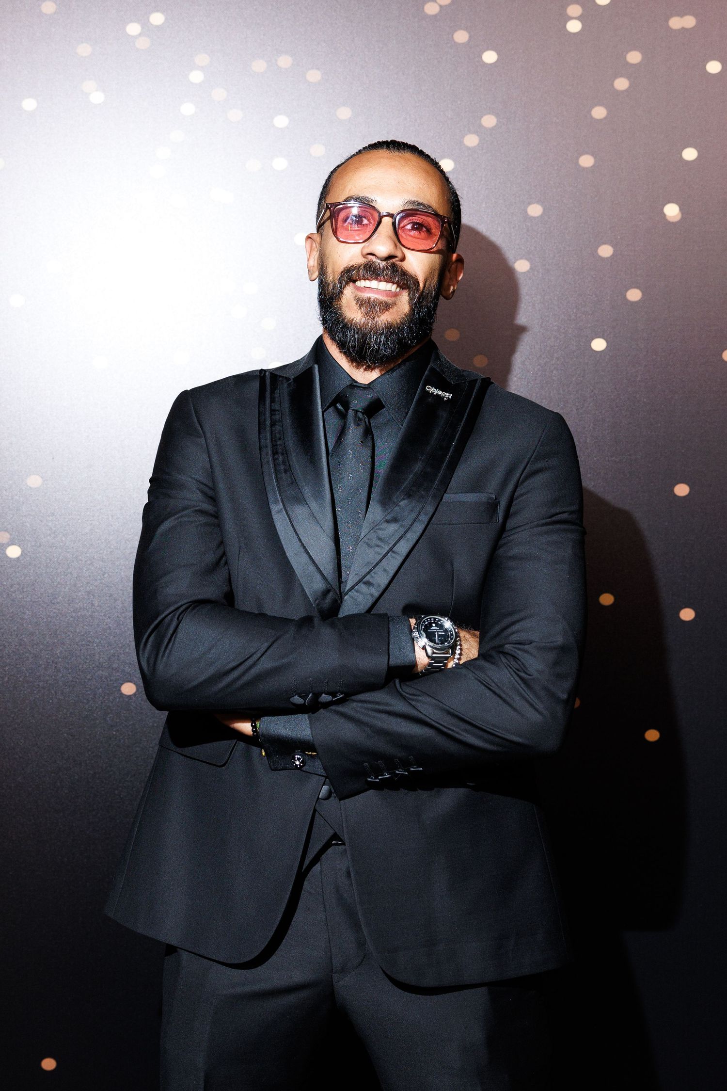 Professional dressed in black formal attire poses against sparkly backdrop at upscale event.