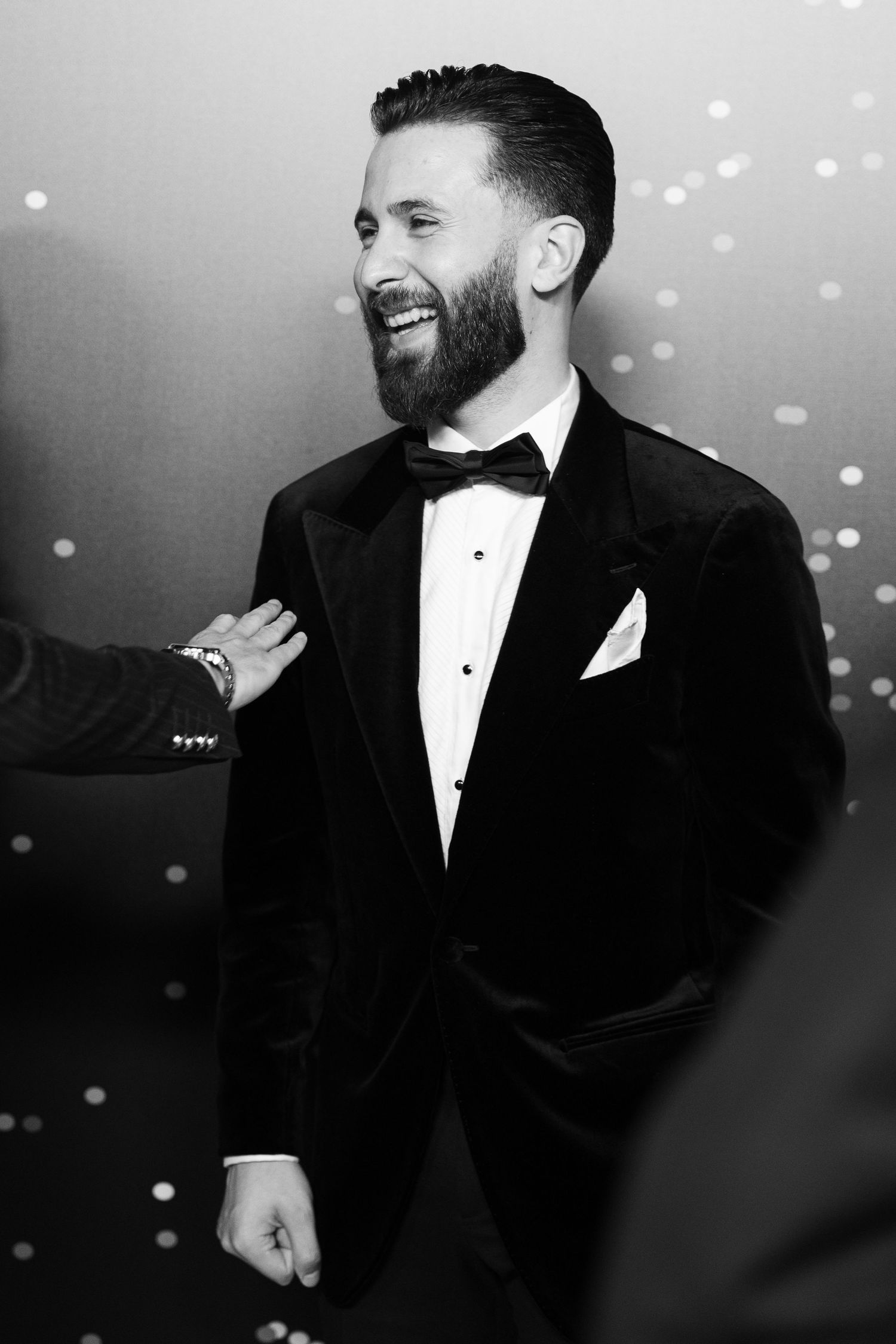Black and white photo of formal event attendees in tuxedos sharing a candid moment.