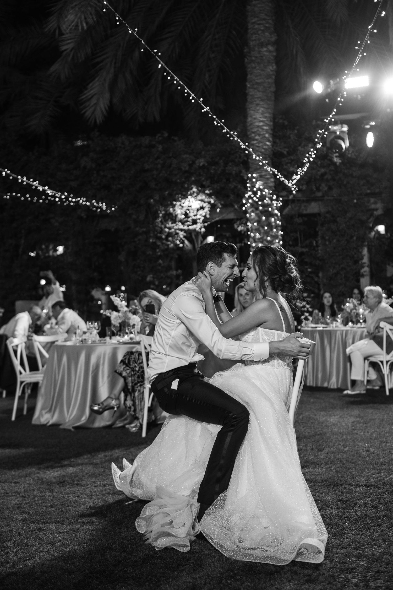 Black and white photo of couple dancing on outdoor lawn under string lights at wedding reception.