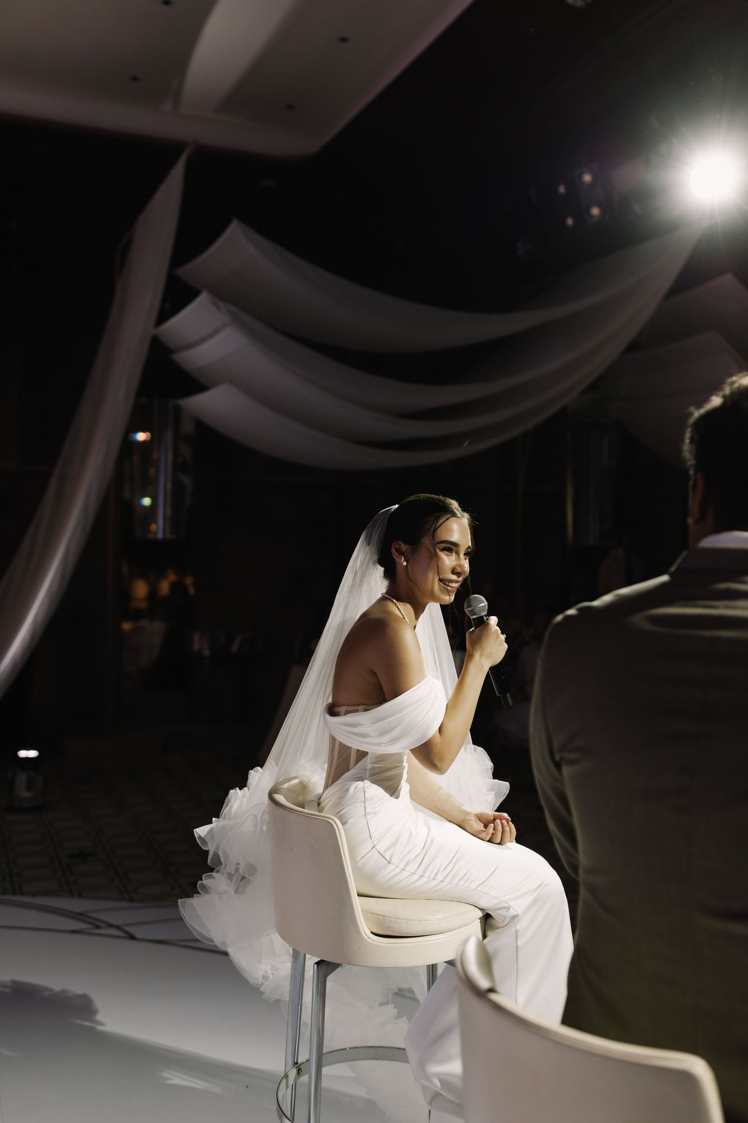 Bride in white dress seated on modern white chair during wedding reception.