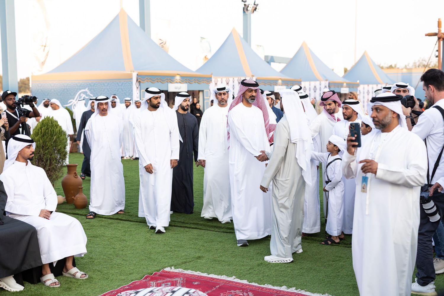 A group of people wearing traditional Middle Eastern white robes gather outdoors under white tent canopies on grass.