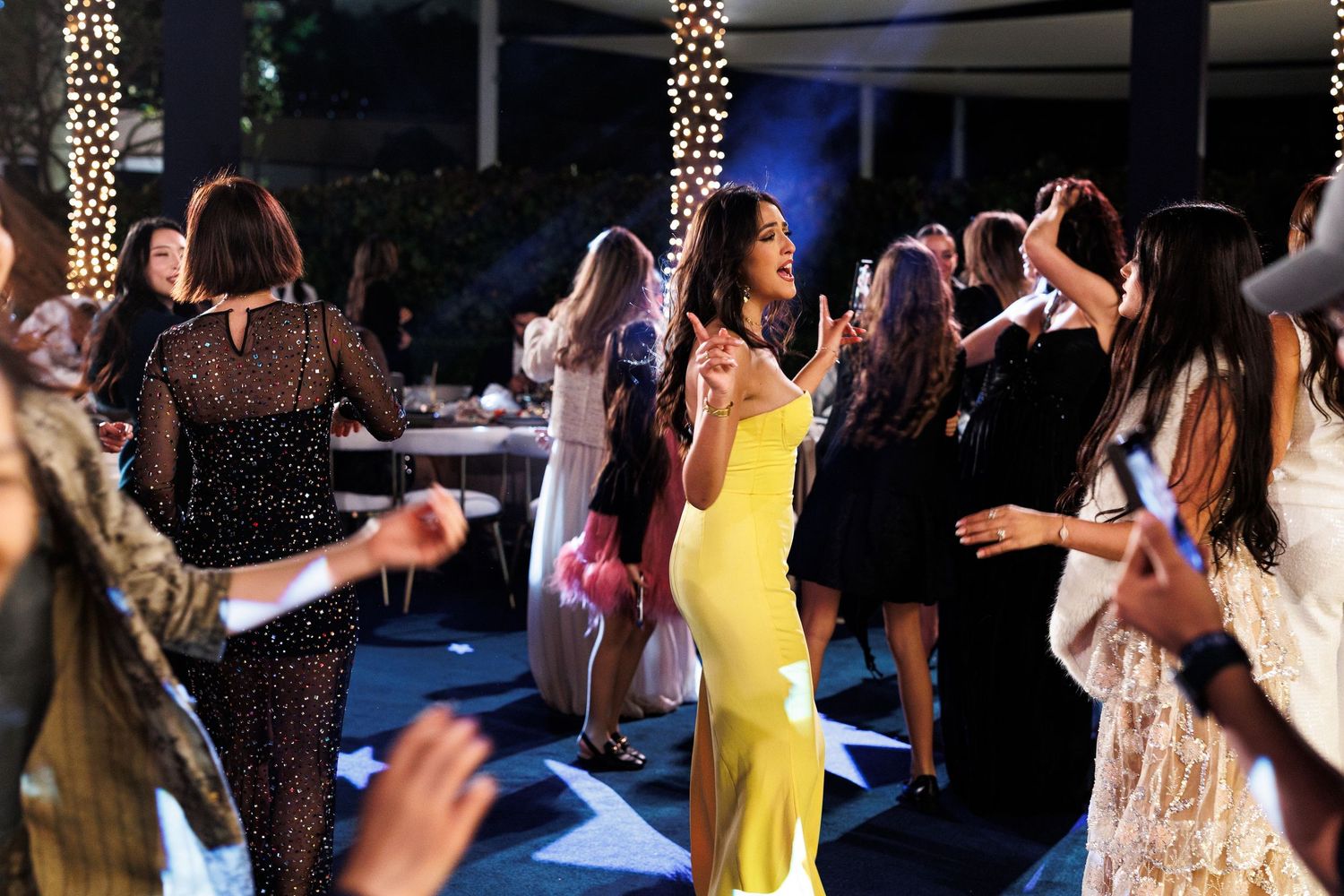 People dancing and celebrating at a festive party with string lights and decorations in the background.