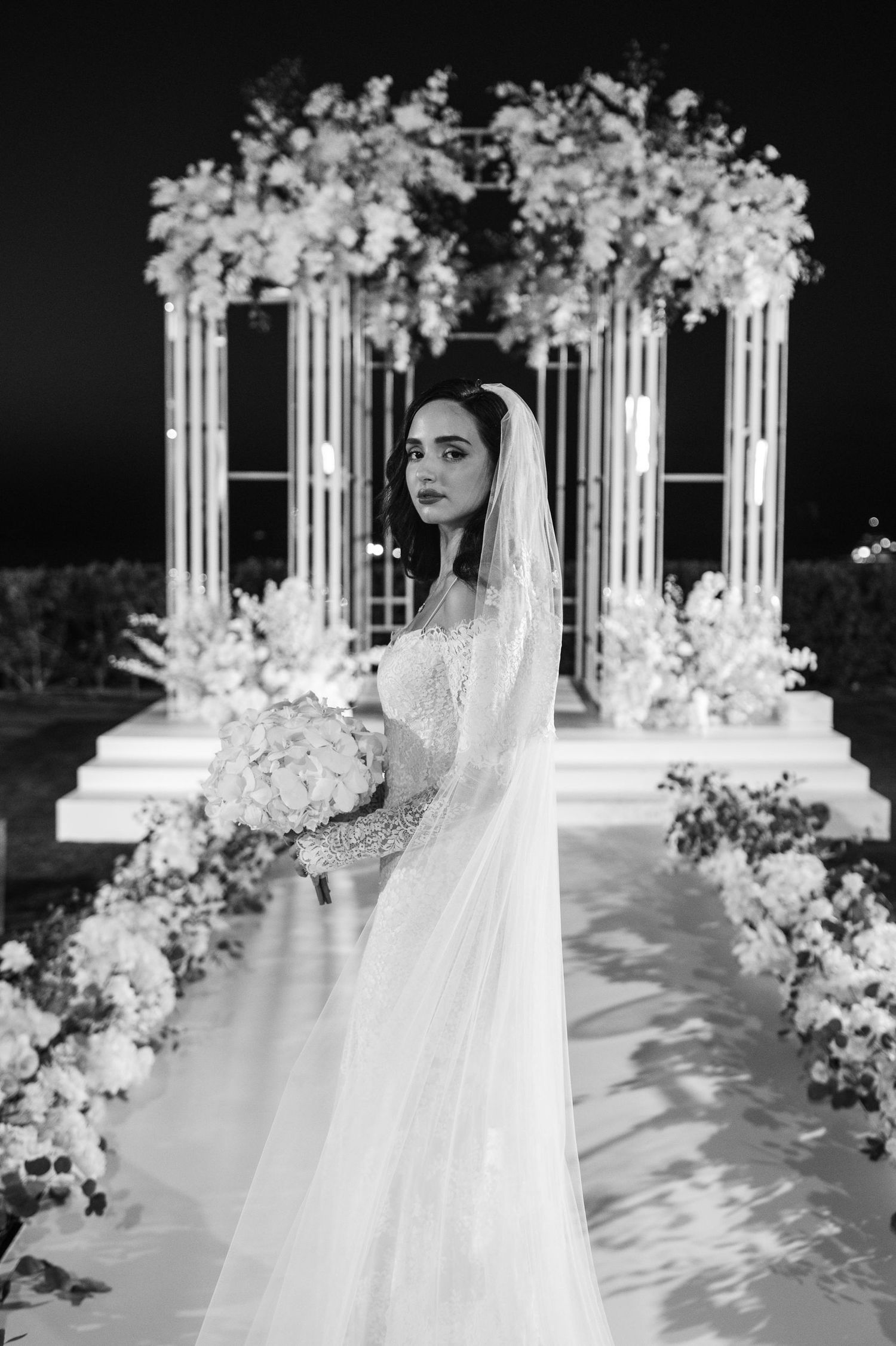 White wedding ceremony setup with decorative columns and floral arrangements against dark backdrop.