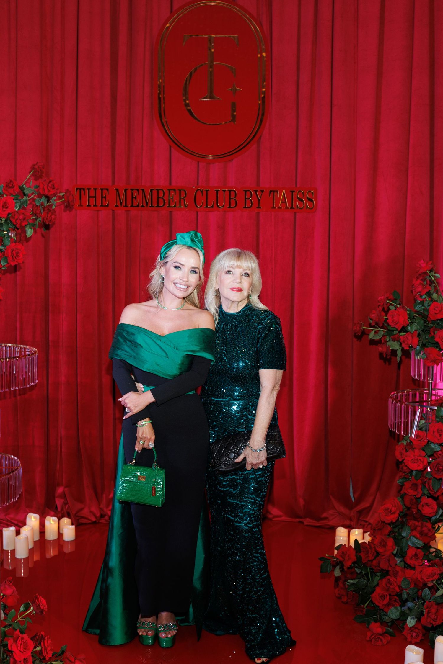 Two guests in elegant evening gowns pose at formal event with red curtain and floral decor.