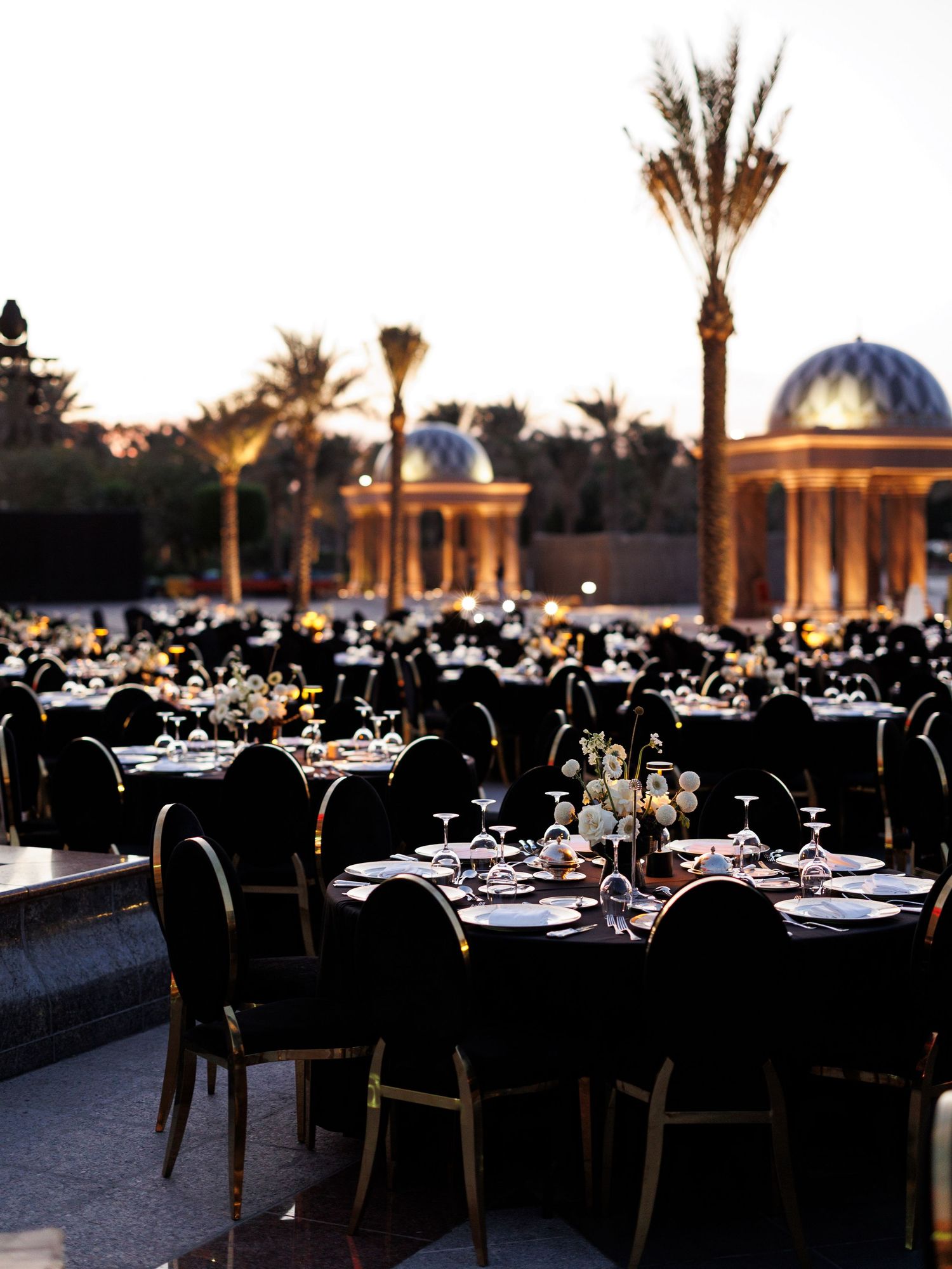 Elegant outdoor dining setup with tables and chairs arranged under palm trees and domed architecture at dusk.
