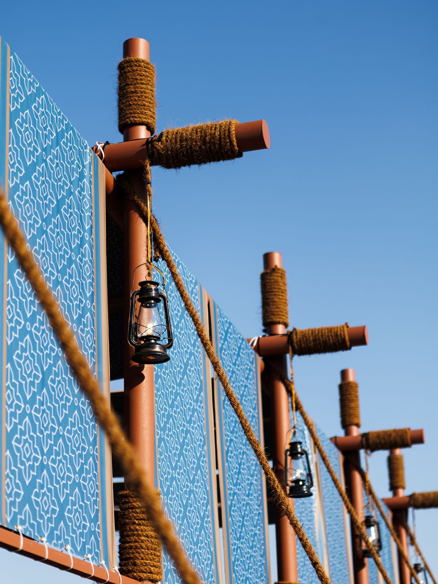 Decorative wooden poles with lanterns and rope wrapping against a bright blue sky create a rustic outdoor display.