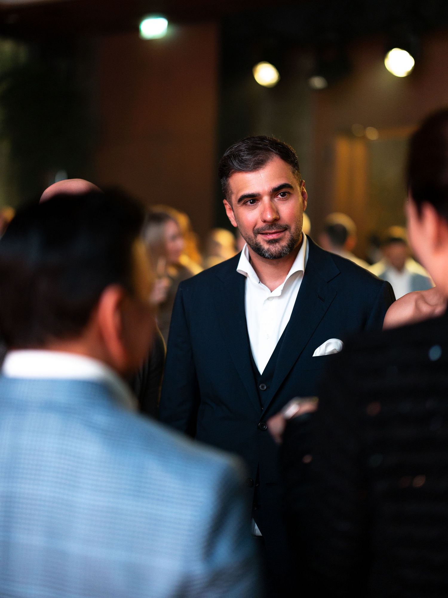 A person in a dark suit and white shirt stands in a dimly lit room with blurred lights in the background.