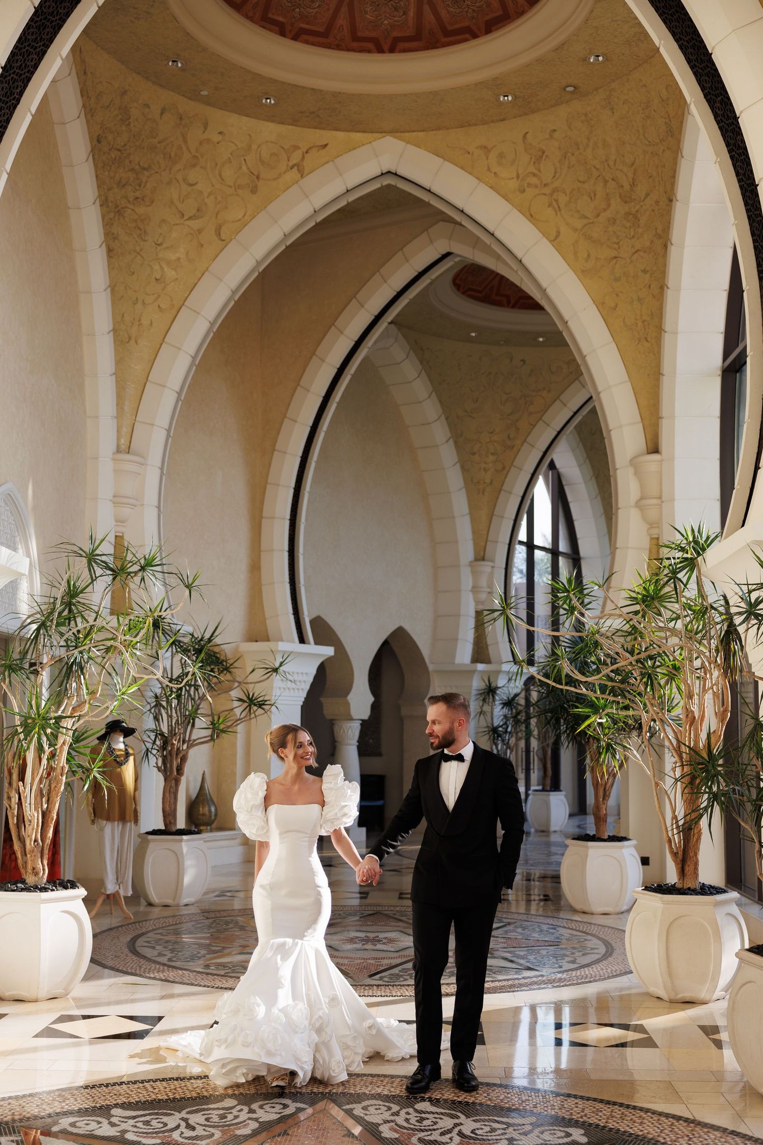 A wedding couple poses in an elegant arched hallway with ornate mosaic floors and potted plants.