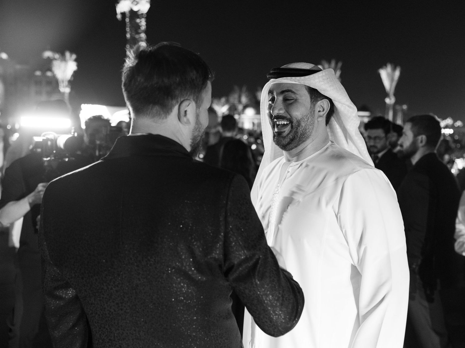 People in traditional Middle Eastern dress and others in suits gather at a dimly lit evening event.