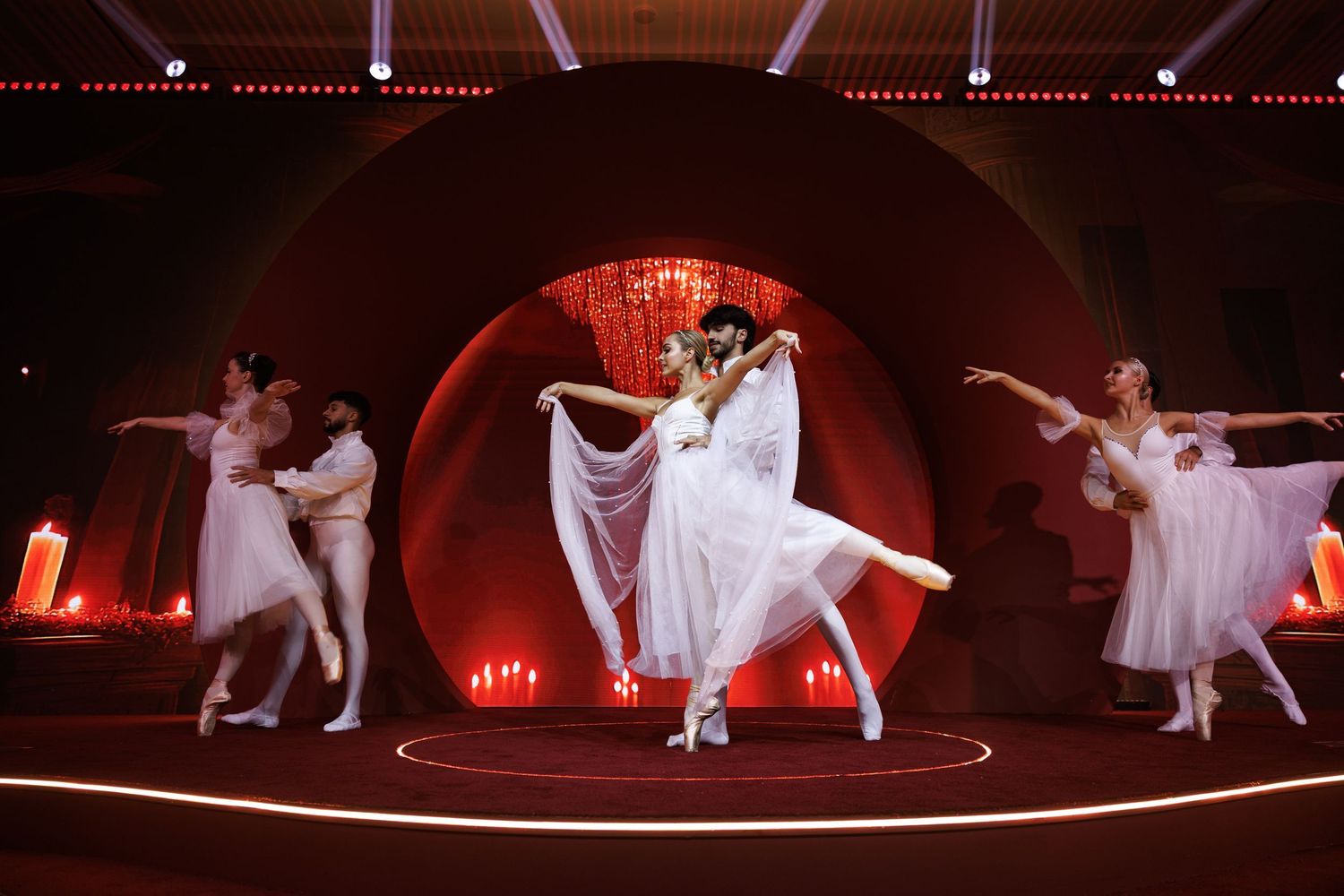 Dancers in flowing white costumes perform dramatic choreography under red stage lighting.