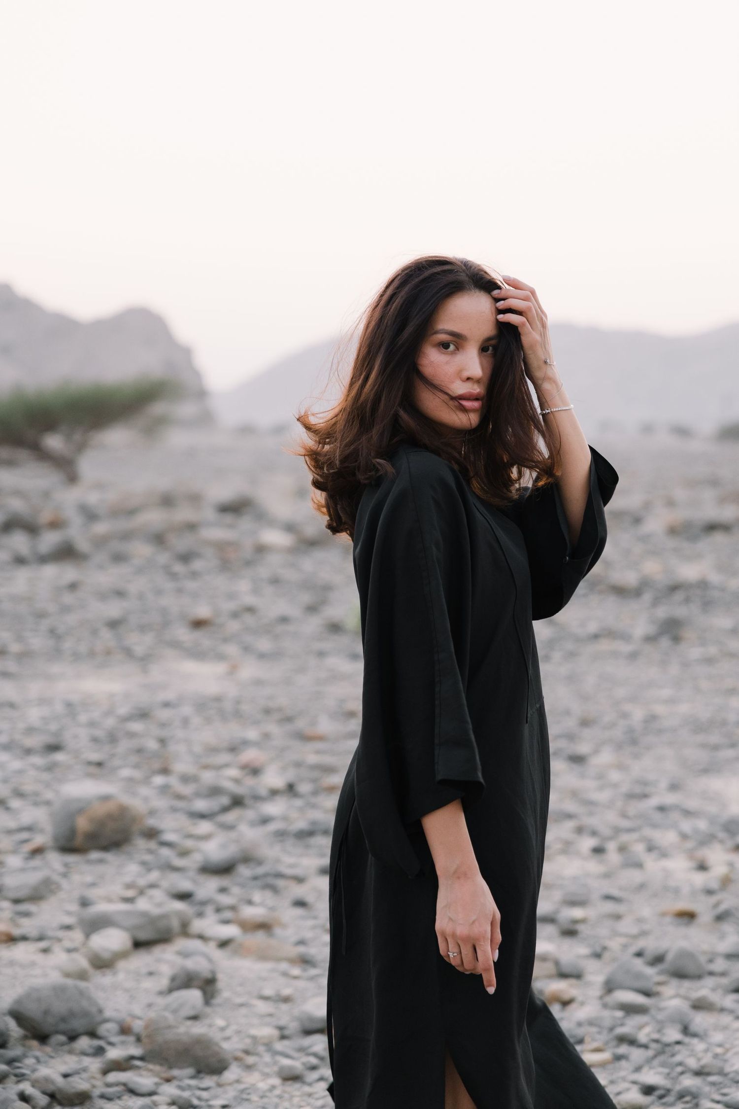 Person in black dress poses dramatically against desert landscape with mountains.