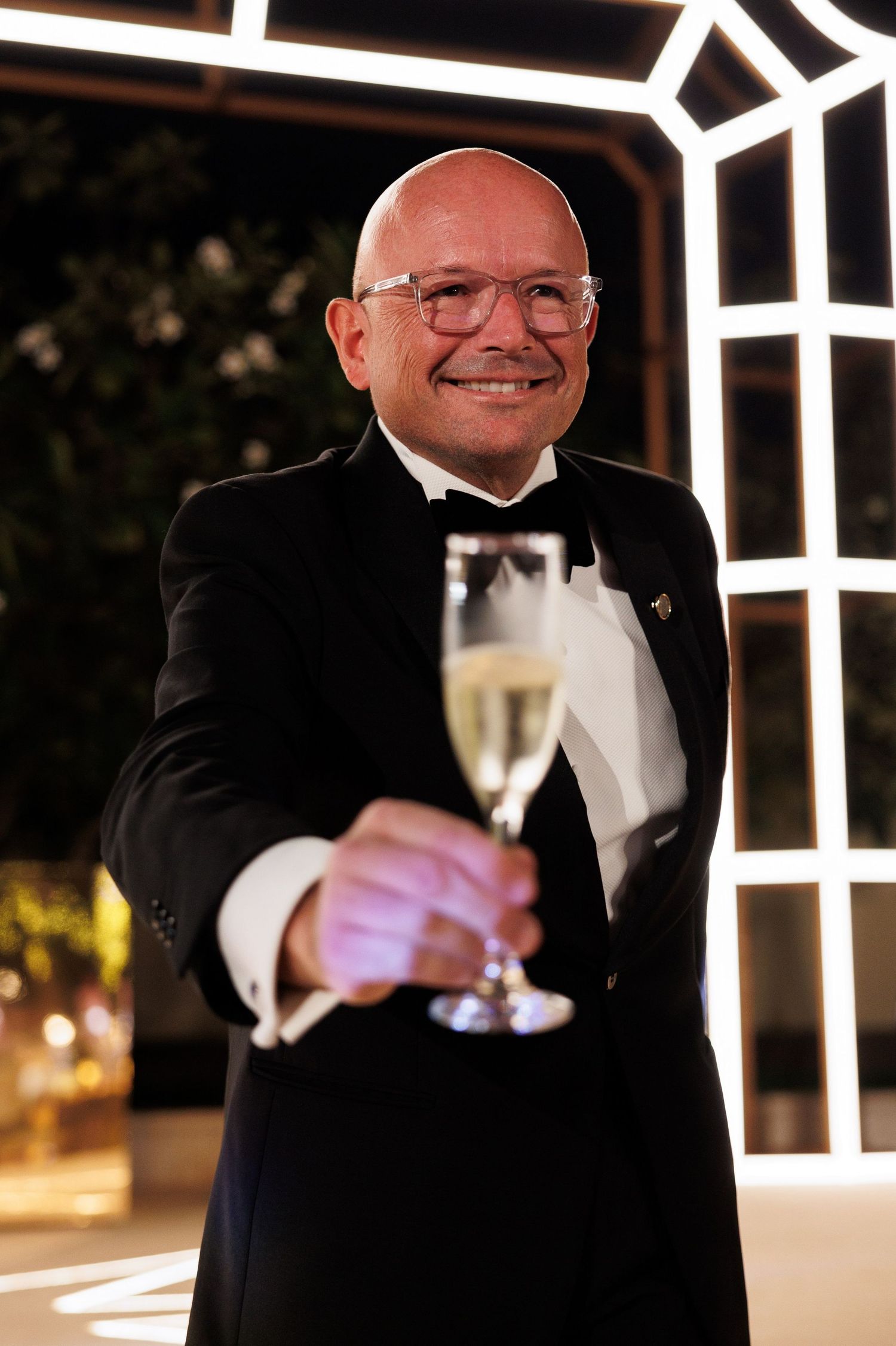 Formal event guest in tuxedo raising champagne glass for celebratory toast.