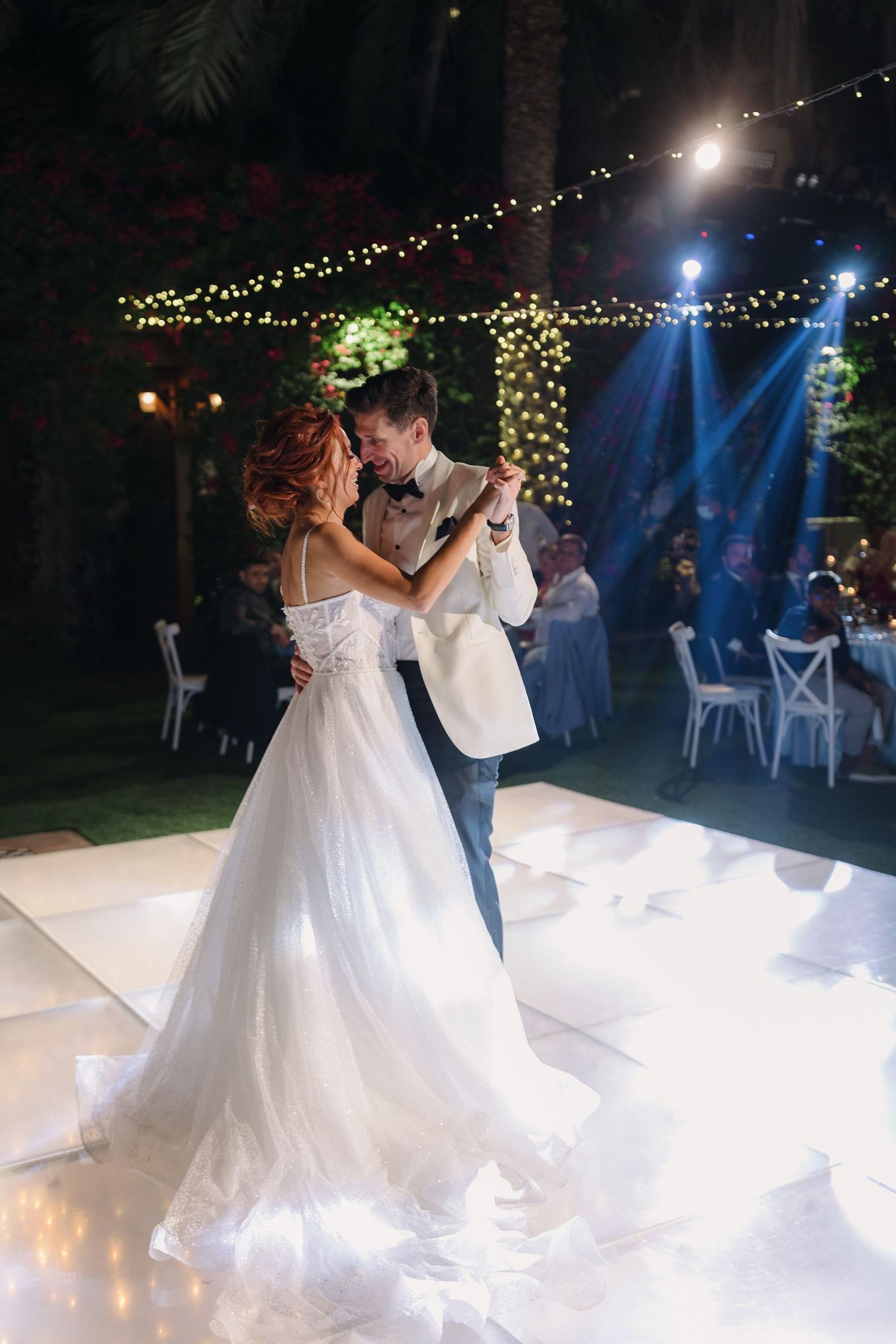 Newlyweds share their first dance under blue spotlights with flowing white wedding dress on outdoor dance floor.