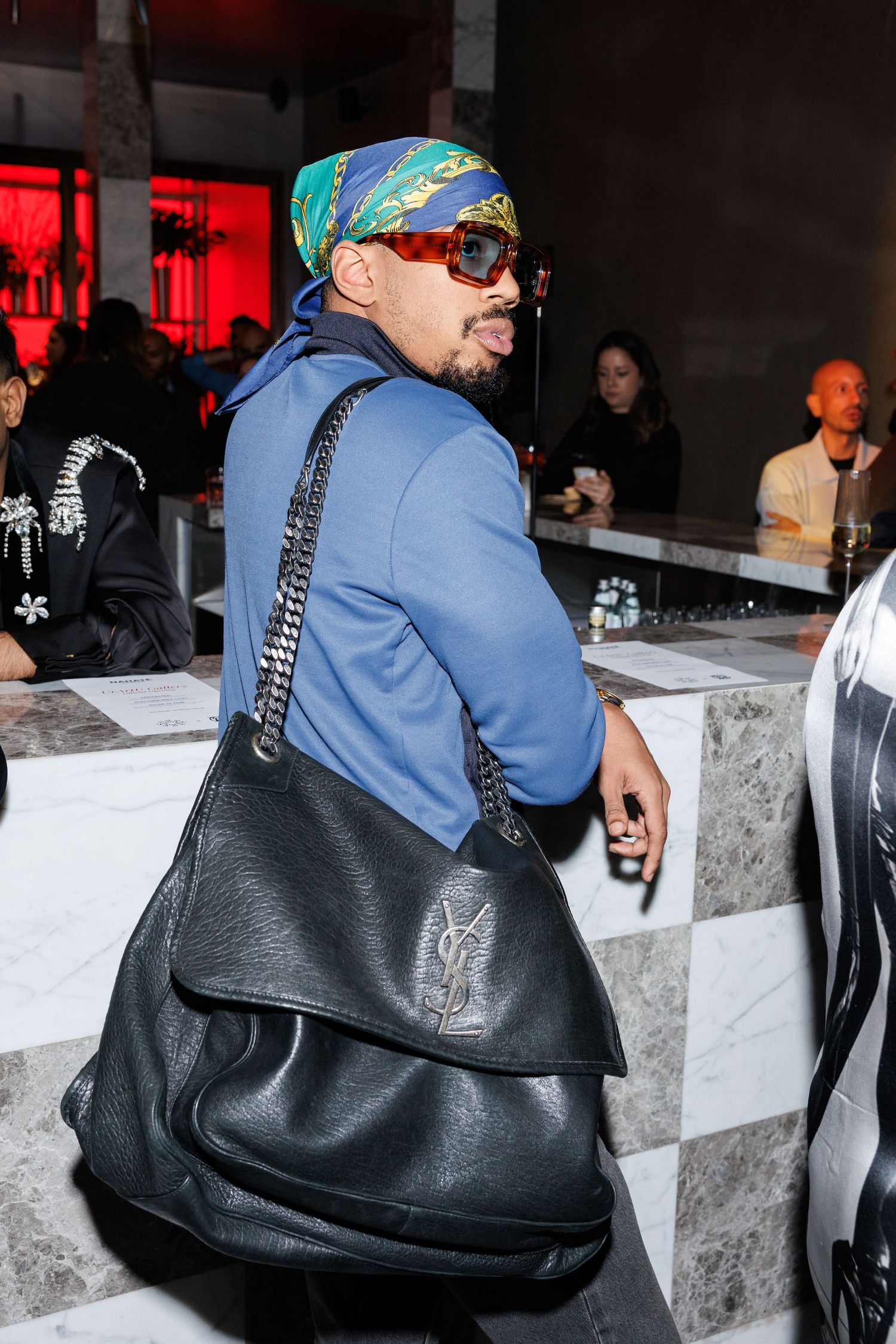 Person in blue jacket and colorful cap wearing sunglasses carries a large black leather YSL bag at a social event.