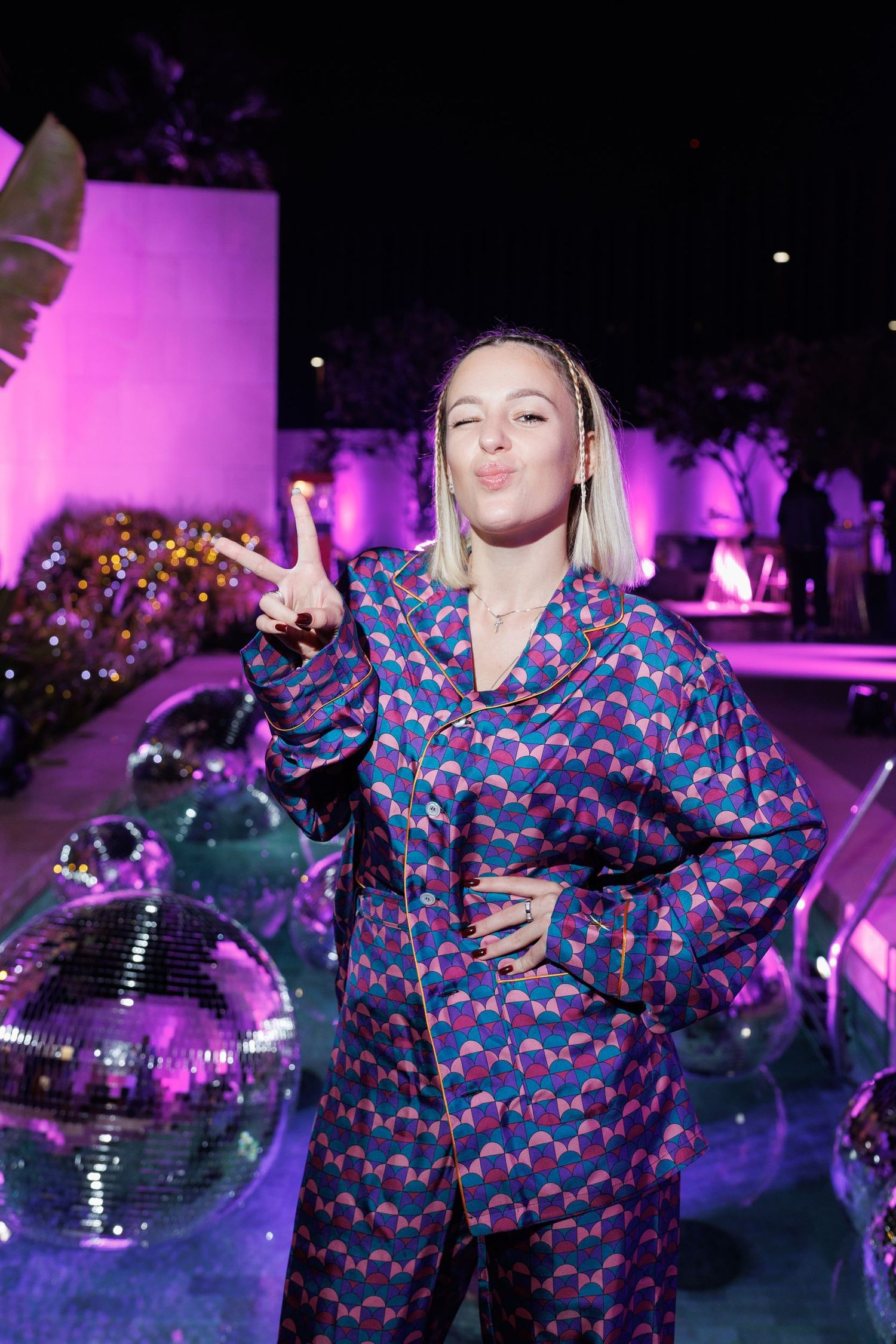 Person in patterned outfit posing with disco ball under purple party lights.
