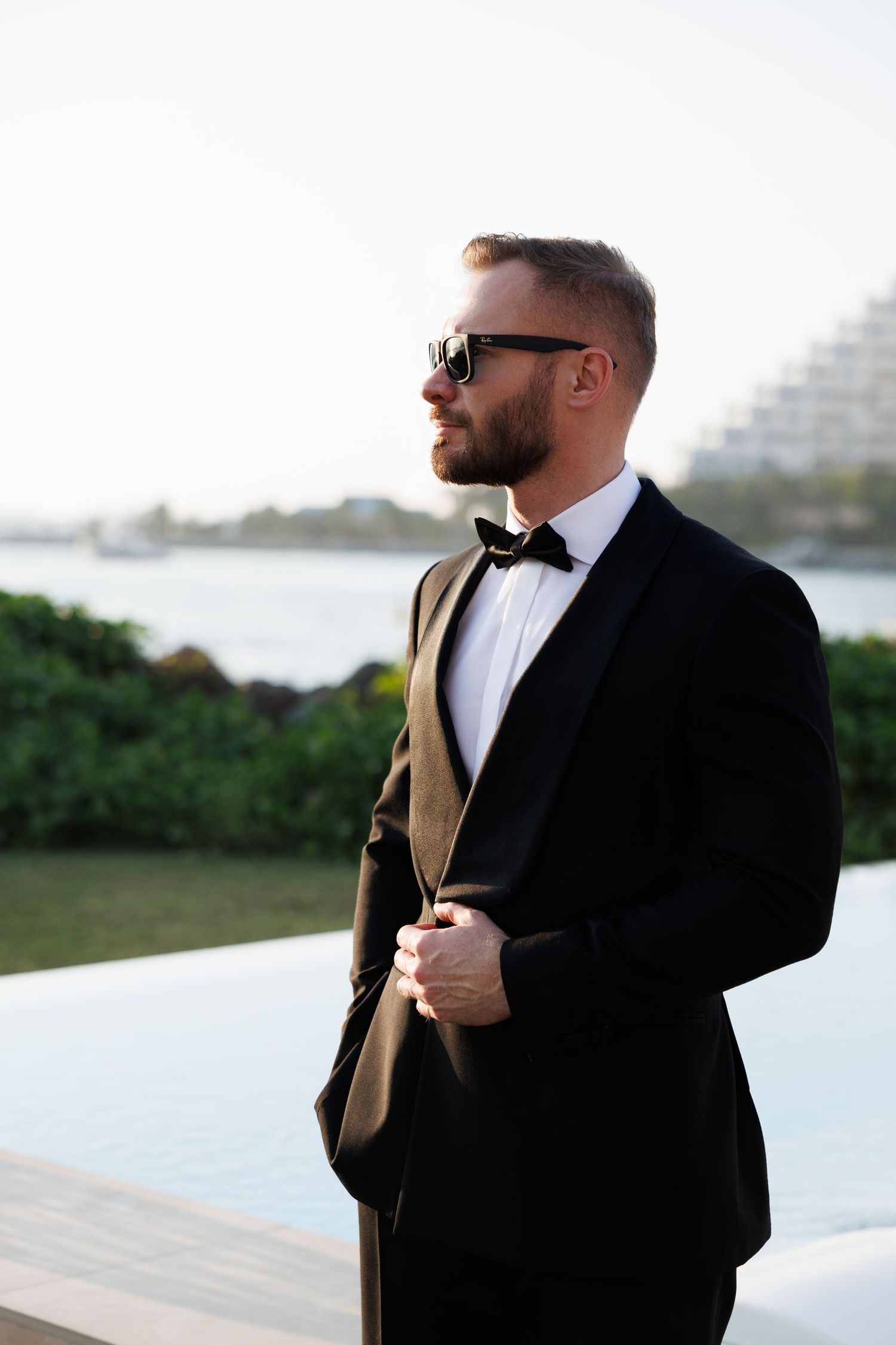 Groom in classic black tuxedo and sunglasses poses against waterfront backdrop.