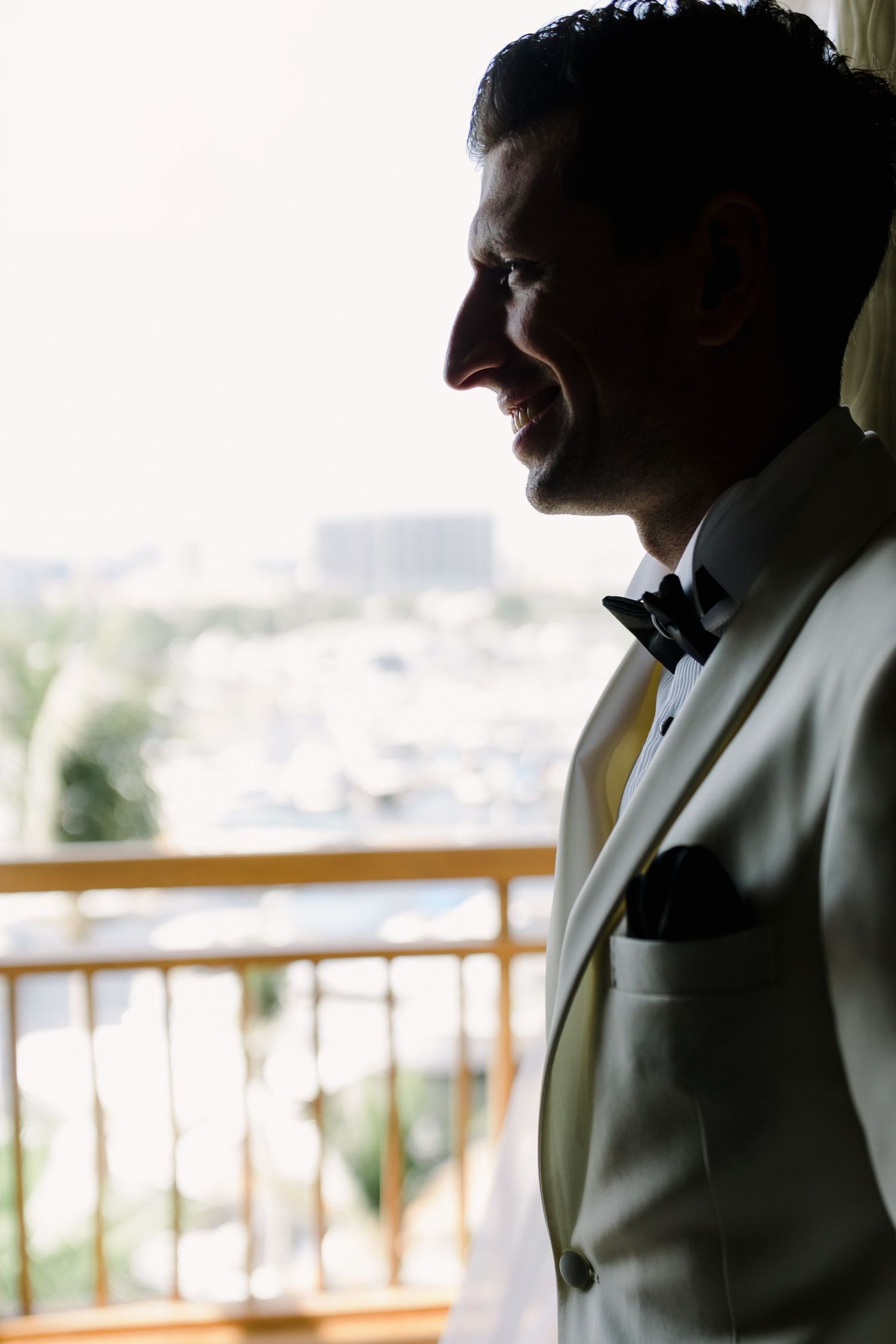 Side profile of a groom in white tuxedo jacket looking out from a balcony with city views.