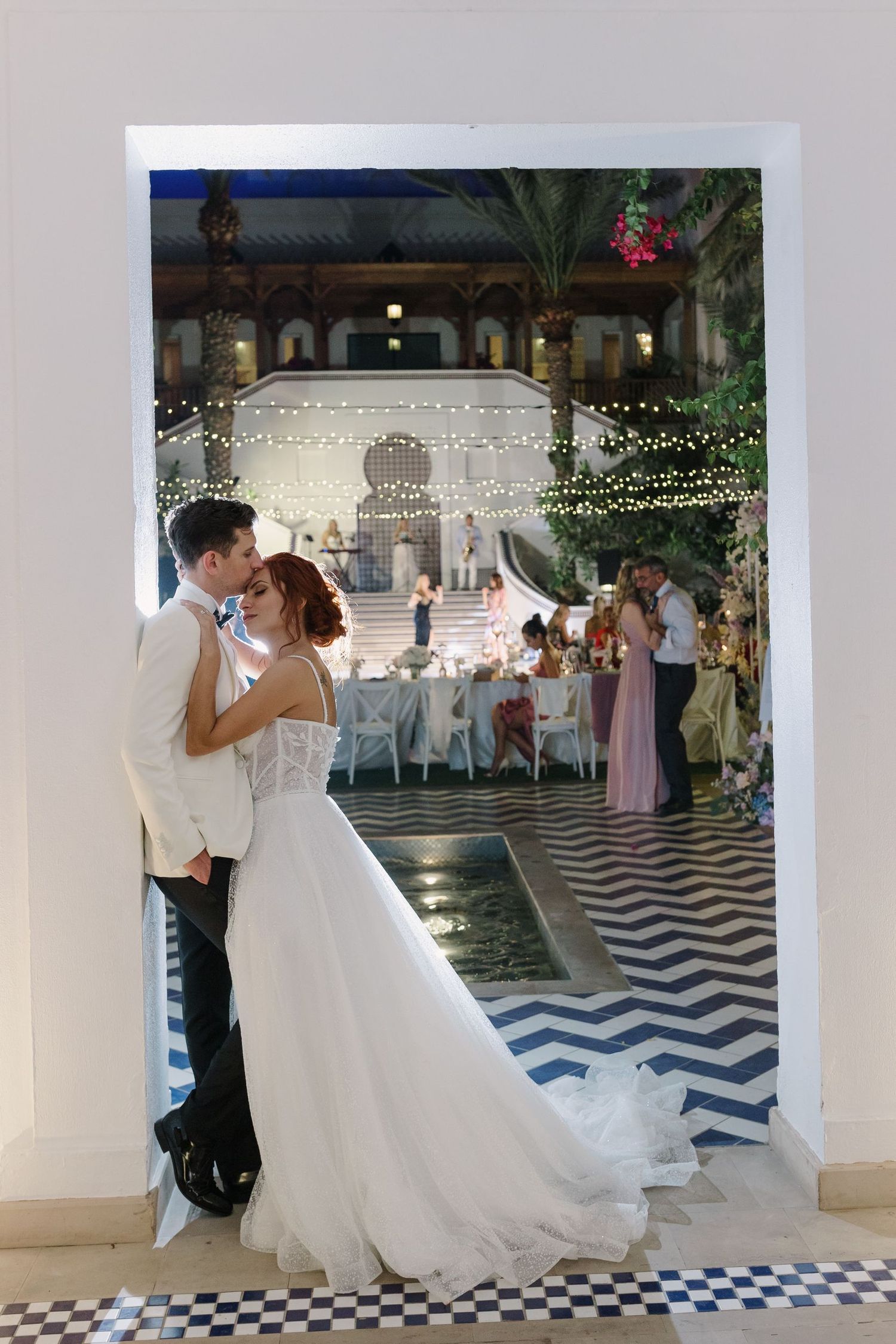 Couple dancing on black and white checkered floor under string lights at wedding reception.